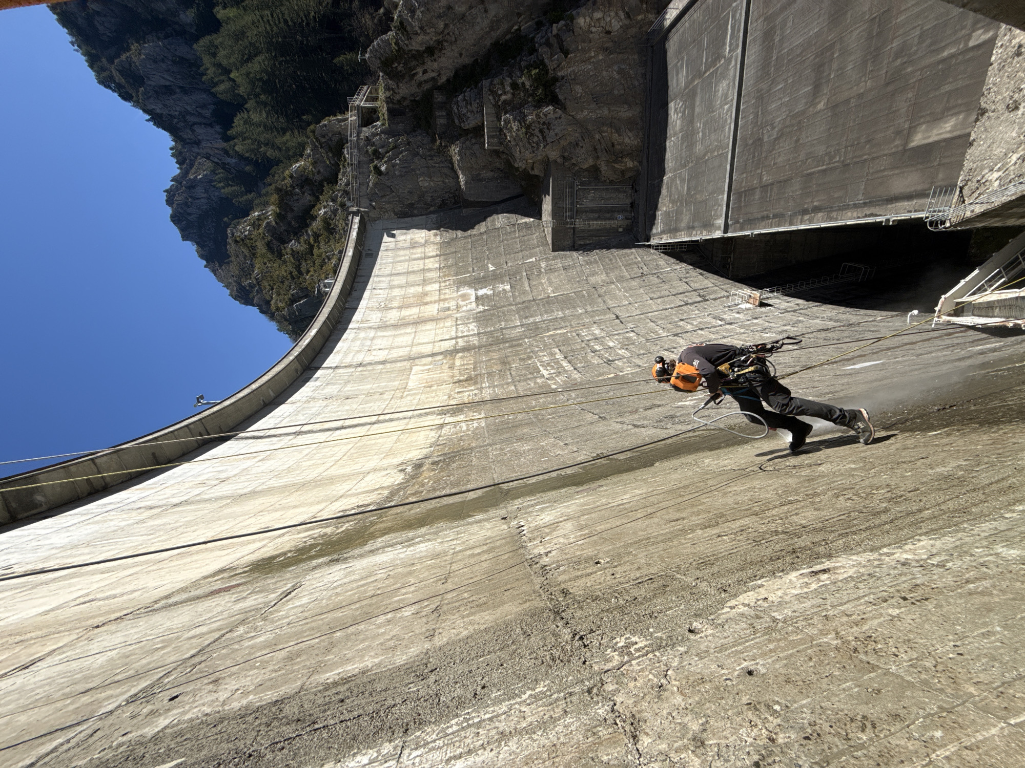 The image depicts a man rappelling down the face of the Castillon Dam in Castellane, France. The dam is a large, curved concrete structure, with the man suspended by ropes from the top. He is wearing safety gear, including a helmet, and appears to be cleaning the dam's surface with a pressure washer. Behind the dam, there is a rocky mountainside covered with trees, under a clear blue sky.