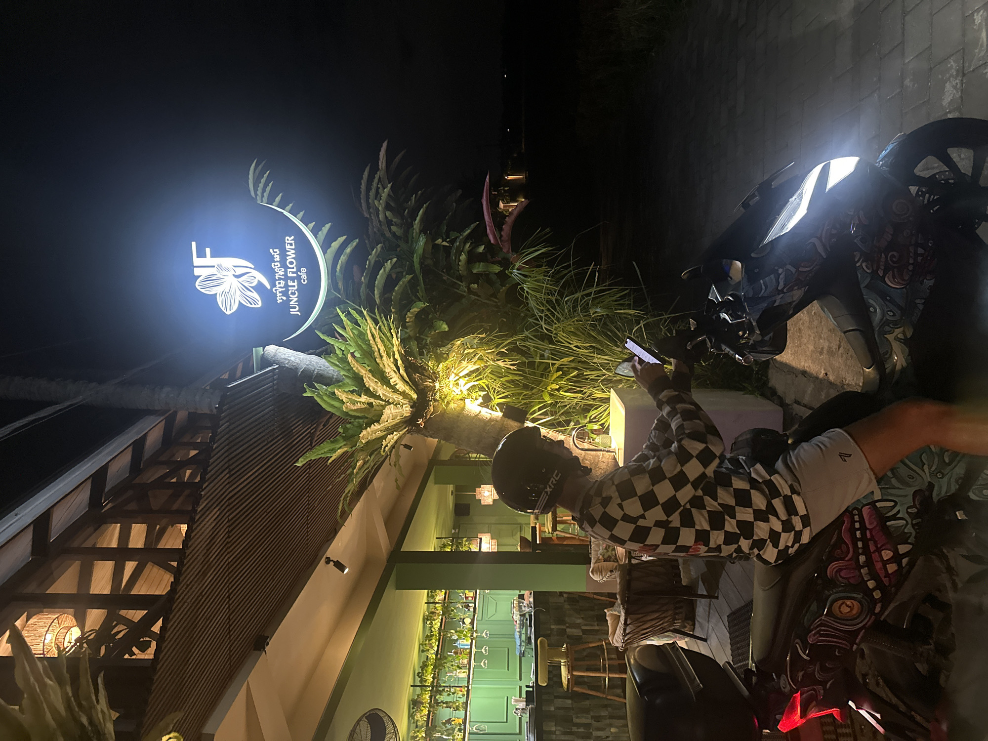 A person wearing a black and white checkered shirt, a black helmet, and gray shorts is sitting on a motorcycle in front of a building with a sign that reads "Jungle Flower cafe." The person is looking at a mobile phone. The motorcycle has a detailed graphic design. The location is in Ubud District, Indonesia. The sign is illuminated and surrounded by greenery. The building is dimly lit, with visible architectural details. Paved brick surfaces are visible in the background.