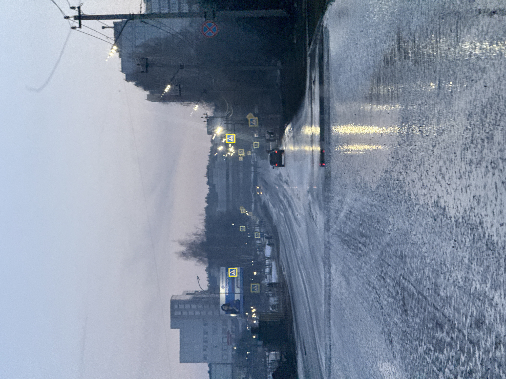 The image shows a rainy or wet street scene in what appears to be the city of Chaikovsky, Russia. The road surface is glistening with water, reflecting the headlights of cars. A dark-colored vehicle is visible on the road. Along the side of the road are buildings, street lights, and pedestrian crossing signs. In the background, there's a hazy or foggy atmosphere. The overall tone is grey and overcast. A power pole is visible in the left top corner of the image.