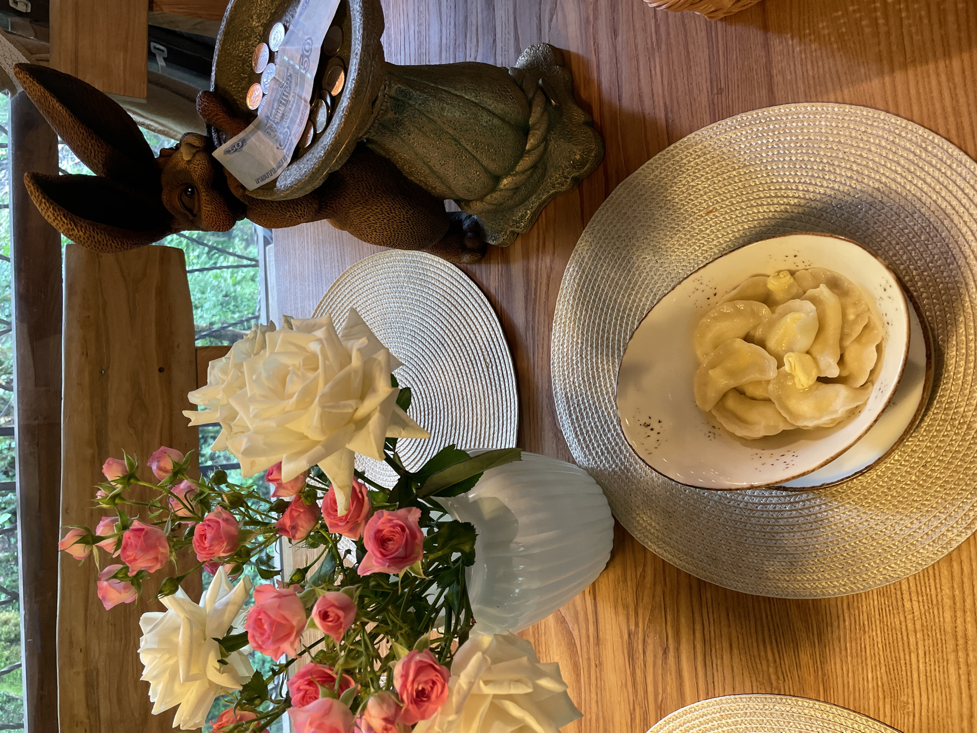 A top-down, indoor shot shows a wooden table arranged with objects. Near the top left, a brown rabbit statue holds a bowl containing coins and a 50 Russian Ruble note. A white vase holds white roses and pink rosebuds, positioned lower left. Two woven placemats rest on the table, one holding a white plate of dumplings topped with butter. The image's location is most likely in Russia, inferred from the currency. No people are directly visible. The focus is on the arrangement of objects on the table. It is not possible to verify the location of Solokh-Aul, Russia, from the image alone.