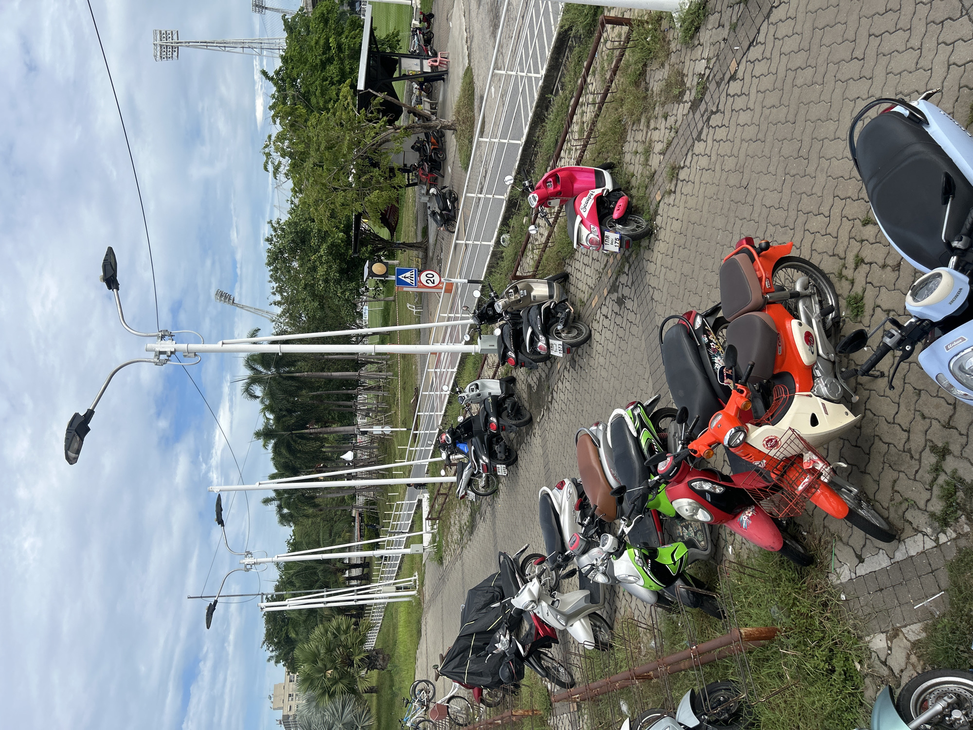 In Tha Khlong Town Municipality, Thailand, a paved parking area is filled with various motorcycles and scooters. The scene is observed from an elevated perspective, looking down on the vehicles and surrounding environment.
Several streetlights, characteristic of urban areas, are visible, some with single lamps and others with multiple. A pedestrian walkway with a white railing runs alongside the parking area, and further in the background, a building with many windows and a stadium with floodlights suggest a public or sports facility. Lush palm trees and other greenery are scattered throughout the landscape, indicating a tropical setting.
A variety of motorcycles and scooters are parked, displaying a range of colors and styles, from classic to modern. Some vehicles are covered with black tarps, while others are exposed to the elements. A few people are visible in the distance, some gathered under a covered area and others walking along the path, suggesting that the location is accessible and possibly a public space. The ground is paved with interlocking gray bricks, and patches of grass grow between the stones and along the edges of the parking area. A traffic sign displaying "20" indicates a speed limit. The sky is partly cloudy, with a mix of blue and white clouds.