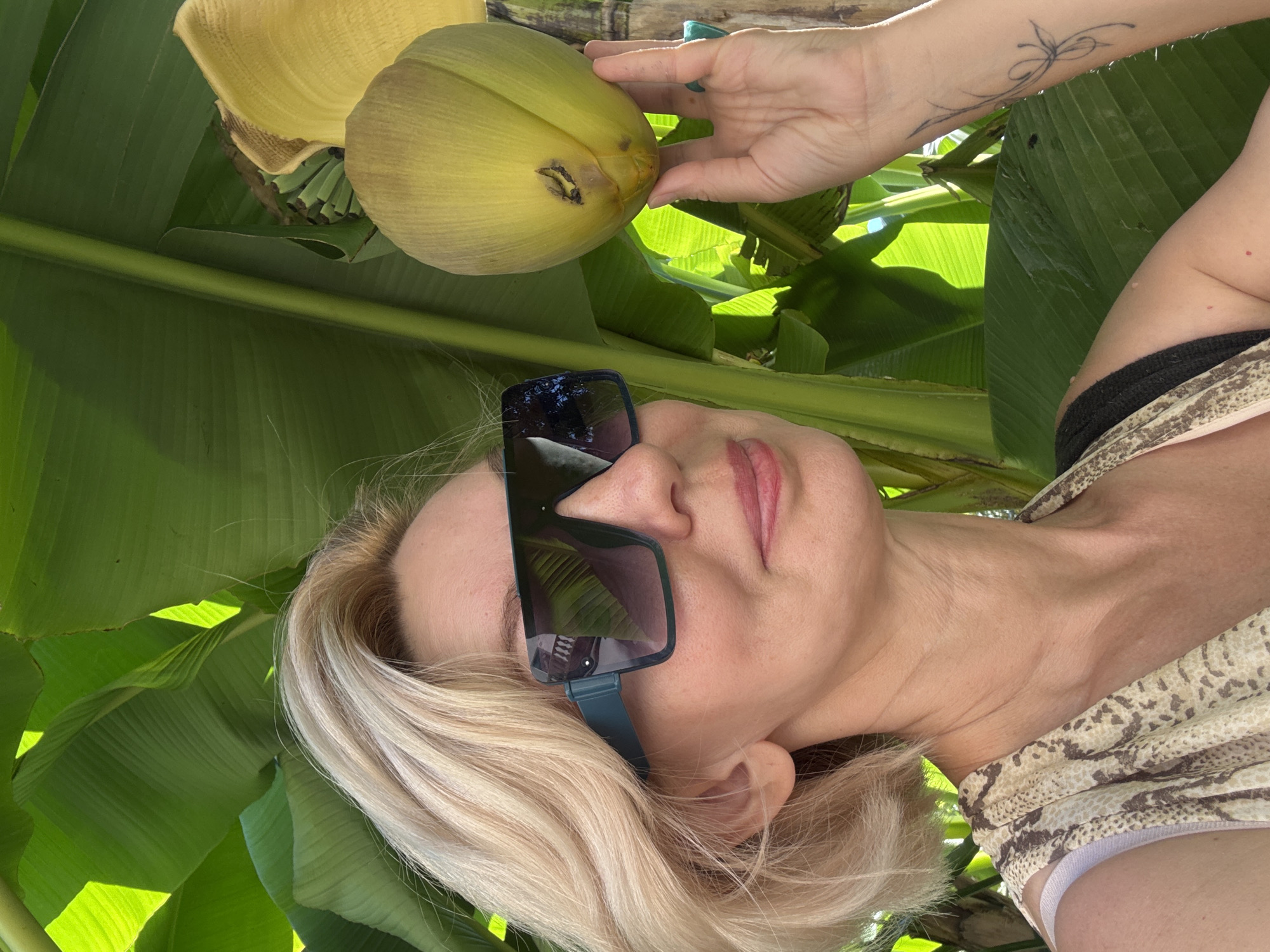 The image shows a woman lying down outdoors, possibly in a tropical setting given the large green banana leaves. She is wearing large, dark-rimmed sunglasses and a patterned top. Her blonde hair is visible around her face. Her arm is raised, and her hand is touching a large, yellow, bulbous object which appears to be a banana flower or a young banana fruit. The lighting suggests it is daytime, with sunlight filtering through the leaves, creating shadows. The woman's facial expression is relaxed, and she appears to be enjoying the moment. There is a subtle tattoo on her forearm. The overall impression is one of leisure and connection with nature.