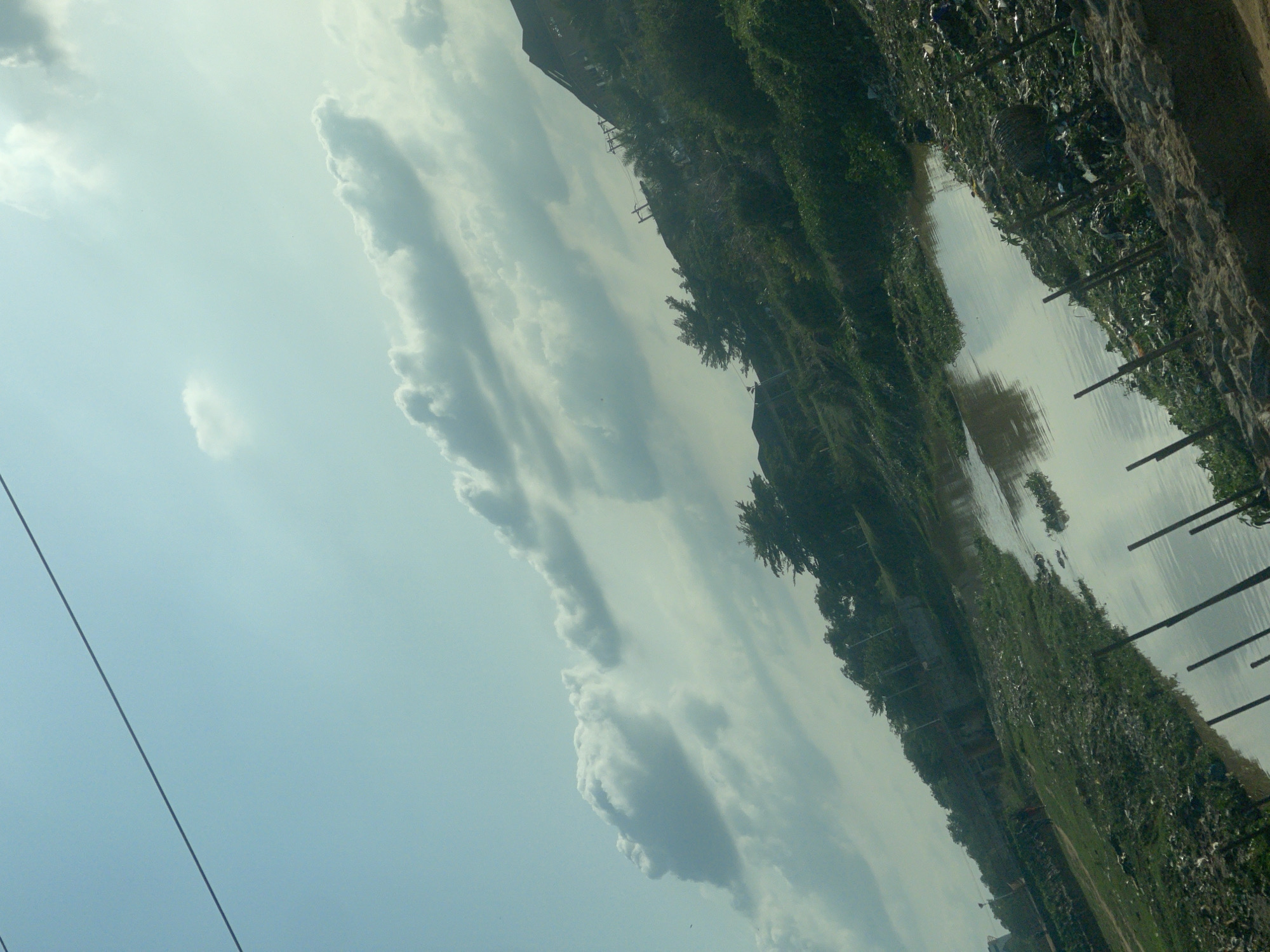The image shows a polluted riverbank in what appears to be a developing area. The water reflects the sky, with visible clouds. The banks are overgrown with vegetation and littered with debris and trash. There are power lines visible against the sky. Utility poles are interspersed along the riverbank, and what appears to be a building can be seen in the distance.
The location in Abuja, Nigeria is inferred due to the evident environmental conditions and infrastructure commonly seen in developing urban areas in that country.