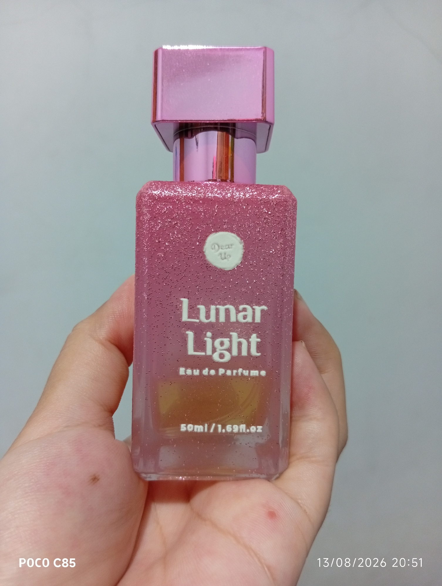 A hand is holding a pink, glitter-covered perfume bottle labeled "Lunar Light Eau de Perfume 50ml/1.69fl.oz". The background is a plain, light blue-grey wall. The image is stamped with "POCO C85" in the bottom left corner and a date and time stamp "13/08/2026 20:51" in the bottom right corner. This image was taken in South Tangerang, Indonesia.