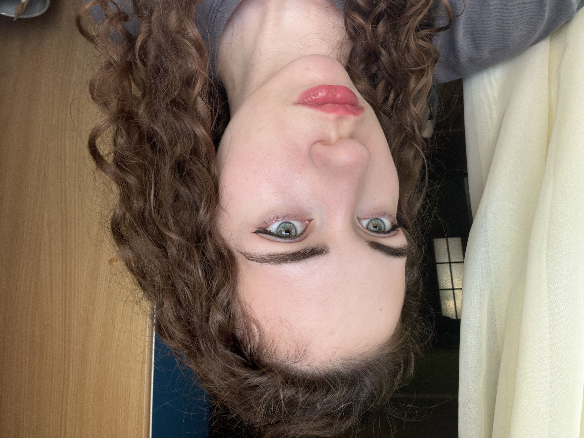 A young woman with curly brown hair and green eyes, wearing a gray t-shirt, is positioned in a head-and-shoulders shot against a backdrop of a sheer cream curtain on the left and a wooden door on the right. She is looking to her left, with her makeup featuring winged eyeliner and a pinkish-red lipstick. The scene appears to be indoors, possibly a room within a residential building. No specific details pointing to Zavodoukovsk, Russia are discernible from the image.
