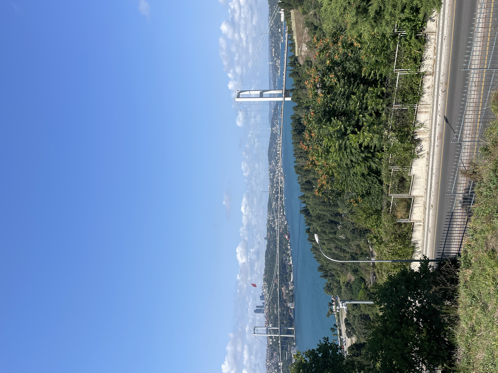 The image captures a panoramic view of the Bosphorus Strait in Üsküdar, Turkey. In the foreground, a lush green hillside slopes down towards a road bordered by a black metal fence. A single street lamp stands on the roadside, its curved metal arm reaching towards the sky. Beyond the road, the vibrant blue waters of the Bosphorus stretch out, dotted with several vessels.
Dominating the middle ground is the majestic Bosphorus Bridge, its white suspension towers rising prominently against the horizon. Cars are visible traversing the bridge, indicating active traffic. The opposite shoreline of the strait is densely populated with buildings, forming a cityscape nestled among rolling green hills. A Turkish flag flies from a pole atop one of the hills on the Asian side. In the distance, several modern skyscrapers pierce the skyline.
The sky is a brilliant, clear blue, with a few scattered white clouds adding texture to the upper portion of the image. The overall scene conveys a sense of urban life intertwined with natural beauty on a sunny day.