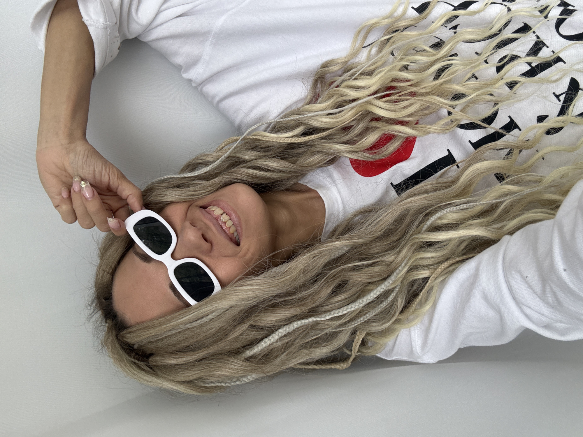 A woman with long blonde hair and white-framed sunglasses is lying down in an indoor setting. She is wearing a white shirt with a graphic design on it. The scene appears to be in a studio or a room with a white background. No location information is available.