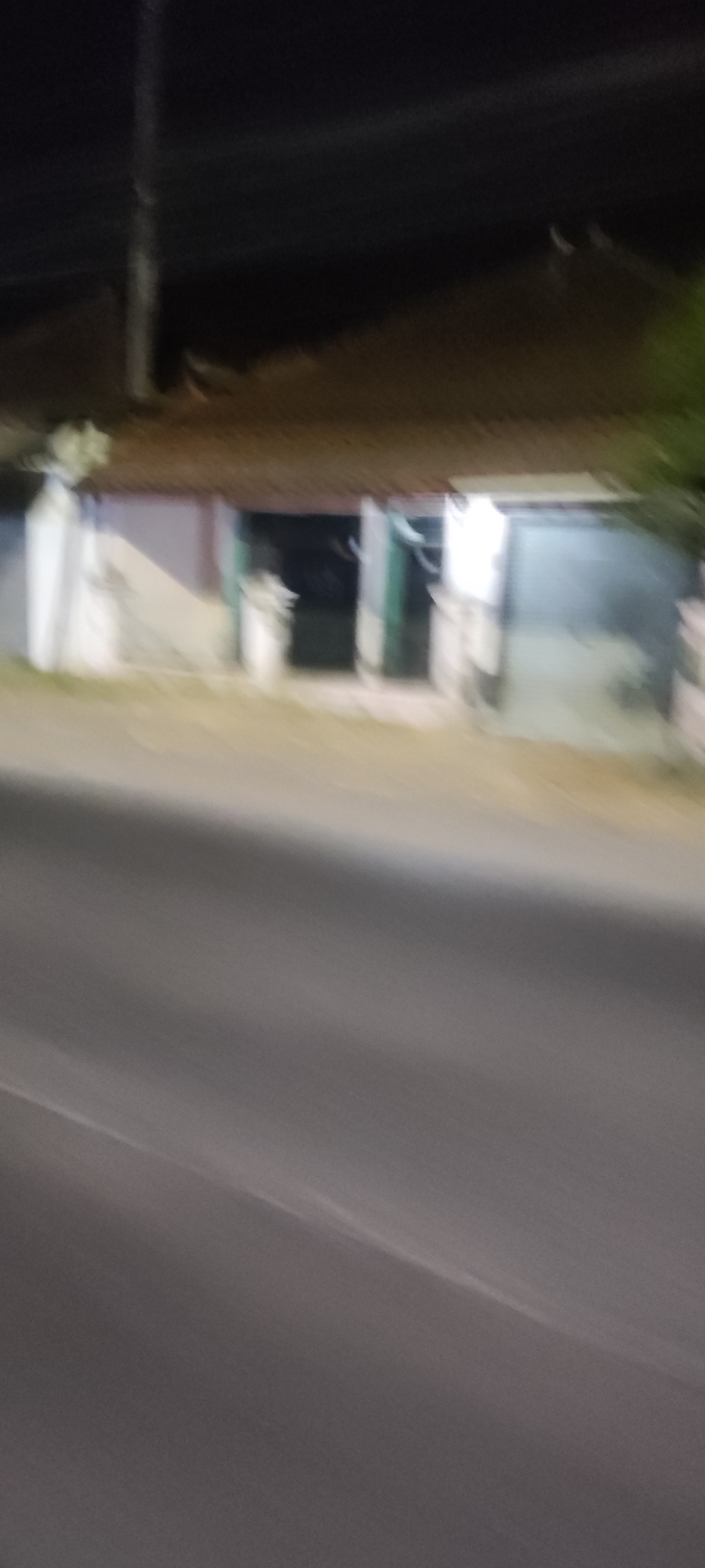 The image is a blurry, night-time photograph that appears to have been taken from a moving vehicle. In the background, there is a low-slung building with a tiled roof. The building has an open entrance and appears to be made of concrete with some white or light-colored sections. There are also some faint green elements, possibly doors or window frames. A dark road occupies the foreground, taking up approximately two-thirds of the image. The overall scene suggests a street view in a residential or commercial area at night. The blurriness of the image makes it difficult to discern specific details about the building's purpose or any objects or individuals that might be present. The location is identified as Babakangebang, Indonesia.