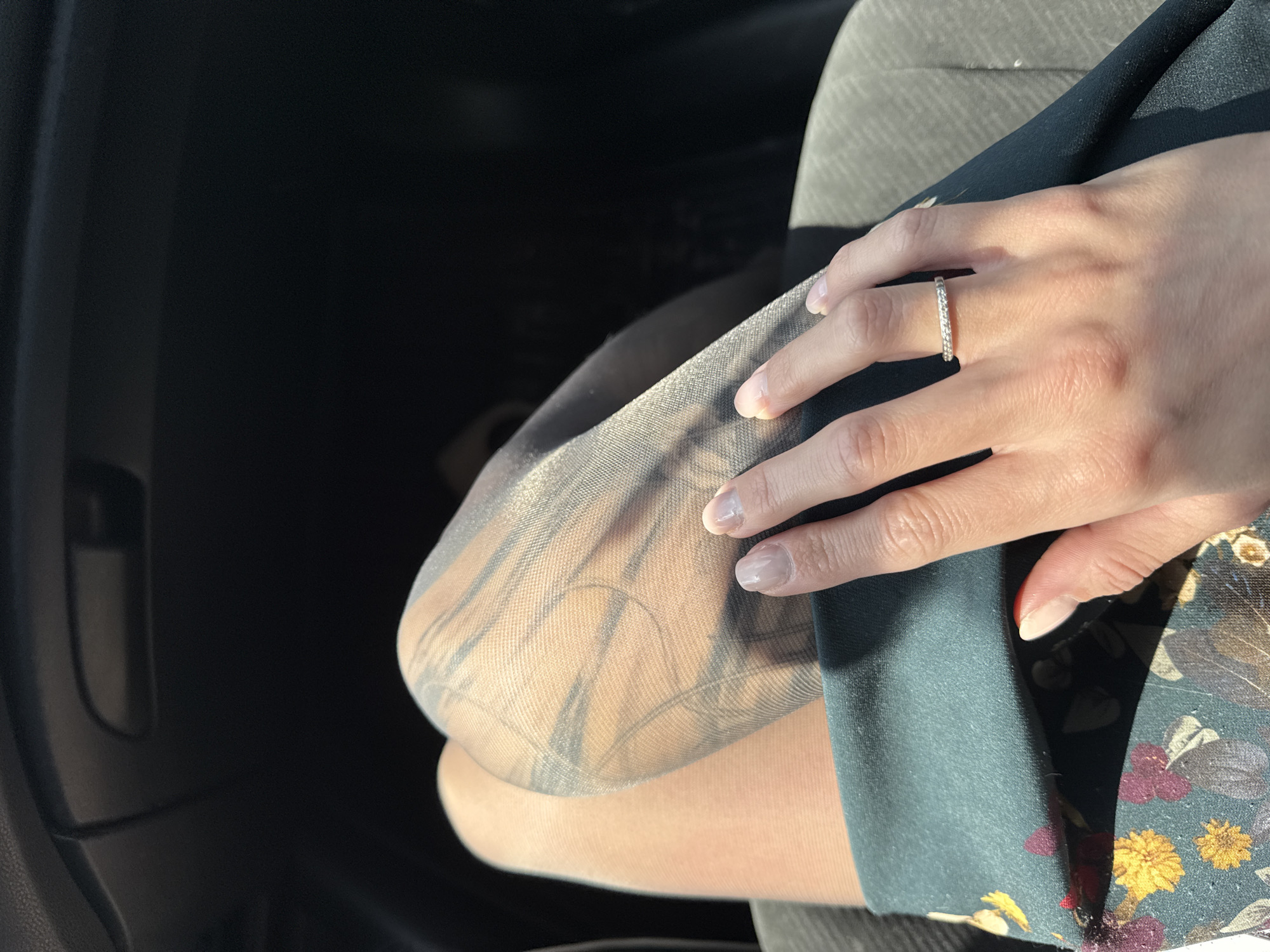 A woman's hand with a diamond eternity ring rests on her knee, which is covered in sheer patterned stockings. The knee is part of a leg crossed over the other, inside a car. A floral-patterned garment is partially visible. The car interior is dark, except for a seat and the leg, which are illuminated by sunlight. The location is unknown and cannot be determined from the image.