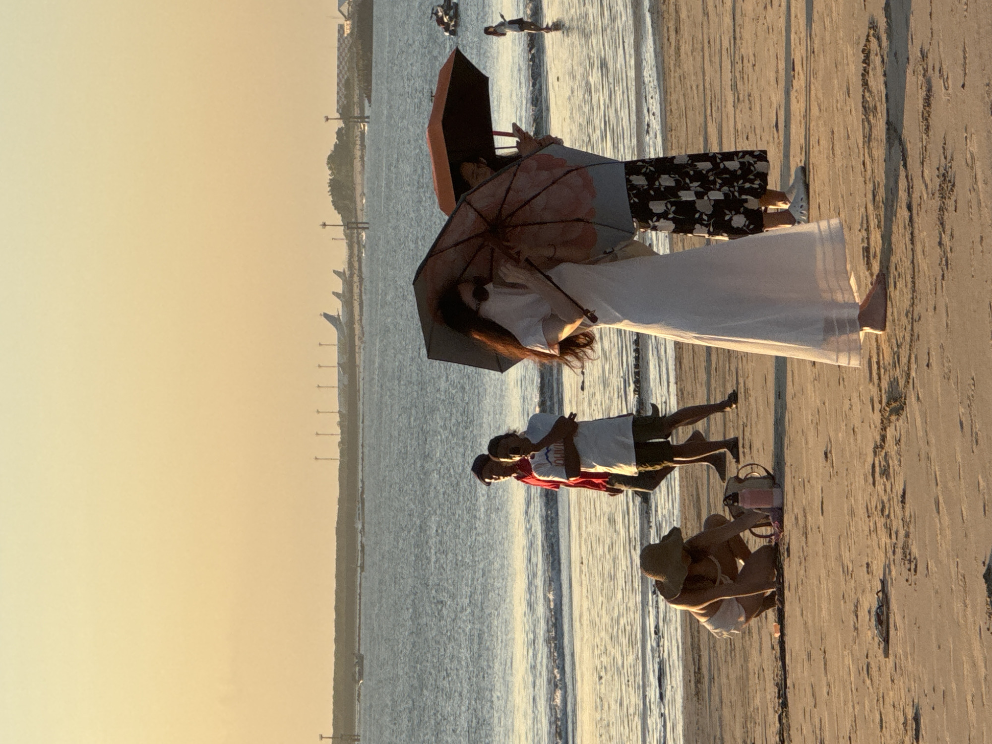 The image depicts a beach scene in Kuta Selatan, Indonesia, during what appears to be sunset or early evening, with a warm, orange-toned sky. In the foreground, a person wearing a straw hat and white shorts is crouched on the sand, interacting with a pink water bottle and a bag. Further back, two individuals walk side-by-side near the water's edge. One is wearing a red shirt and dark shorts, while the other wears a white t-shirt and khaki shorts. In the midground, two women are standing, each holding an umbrella. The woman on the left wears a white dress, and the woman on the right wears a black skirt with a floral pattern. In the background, the ocean stretches out to the horizon, with a distant shoreline featuring trees and structures. Several airplanes are visible on the tarmac of an airport in the distance, and a few boats are on the water. The overall atmosphere is relaxed and summery.