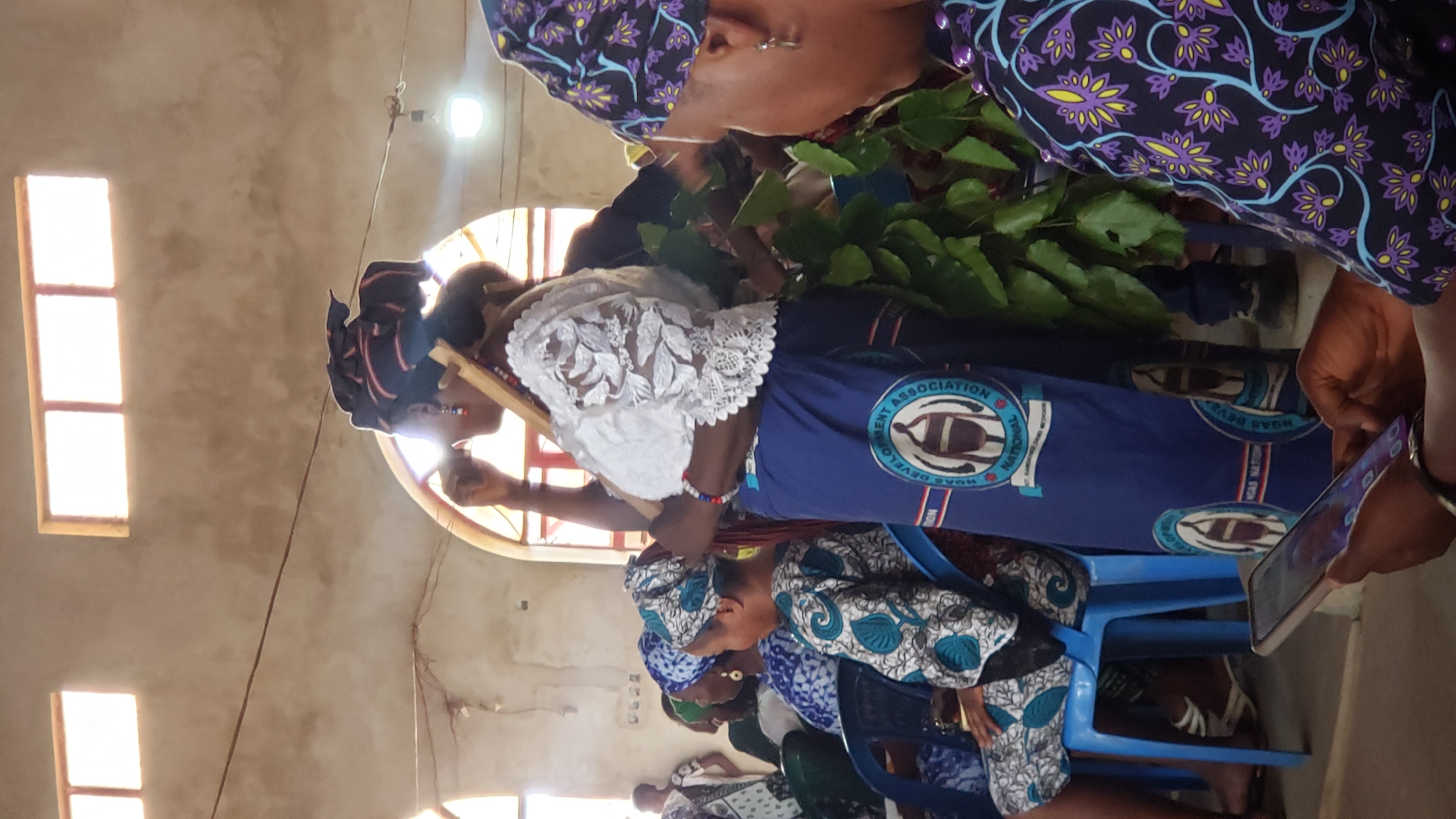 The image depicts a group of people, possibly in Narayi, Nigeria, gathered indoors. Some are seated on blue plastic chairs, while others stand. Several individuals wear clothing with a blue and white pattern, possibly a uniform or attire for a specific association, as evidenced by a logo including the text "HOAS DEVELOPMENT ASSOCIATION". One standing woman is wearing a white lace top and has a headwrap. She's holding what appears to be a wooden frame or stick. Another person appears to be taking a picture or video with a cell phone. There are plants or foliage visible, potentially being used in some sort of ceremony or event. Light enters the room through windows, illuminating the scene. The walls are a muted beige color.