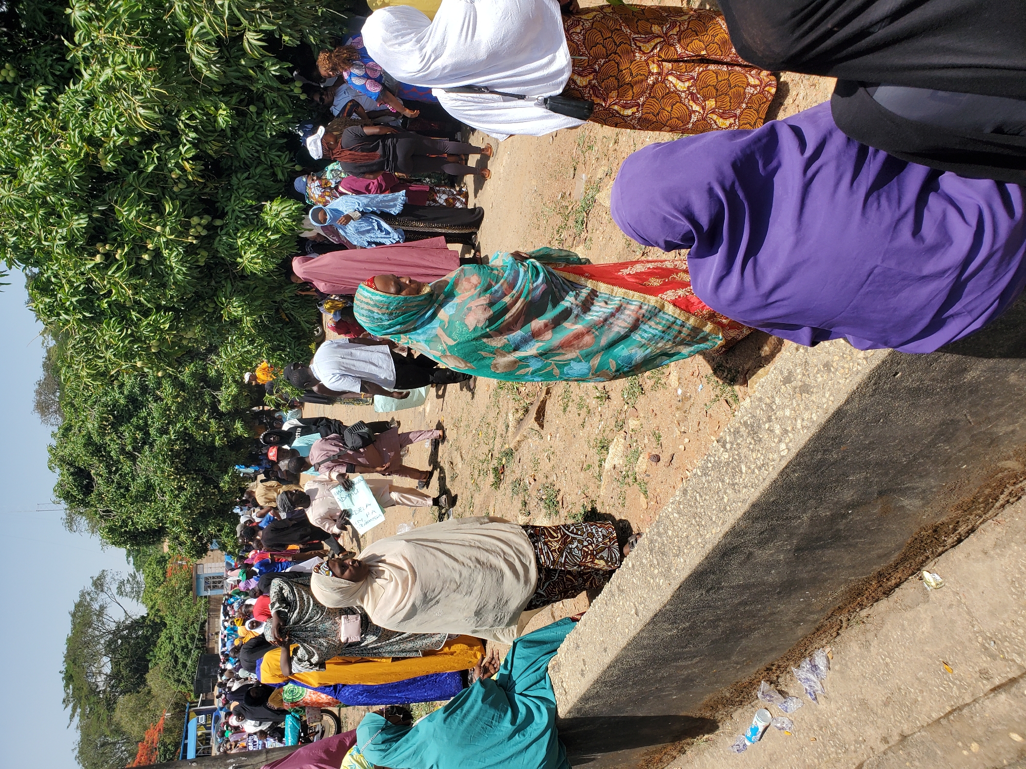 A large group of people, predominantly women, are gathered outdoors in Jos, Nigeria. Many are wearing traditional Muslim attire, including headscarves of various colors such as white, purple, green, and black. The crowd appears to be either waiting in line or protesting. Some individuals are holding signs with text in a local language. The surrounding environment includes trees, buildings, and concrete structures. The ground appears to be dirt or gravel. There is litter along the edge of a concrete barrier.
