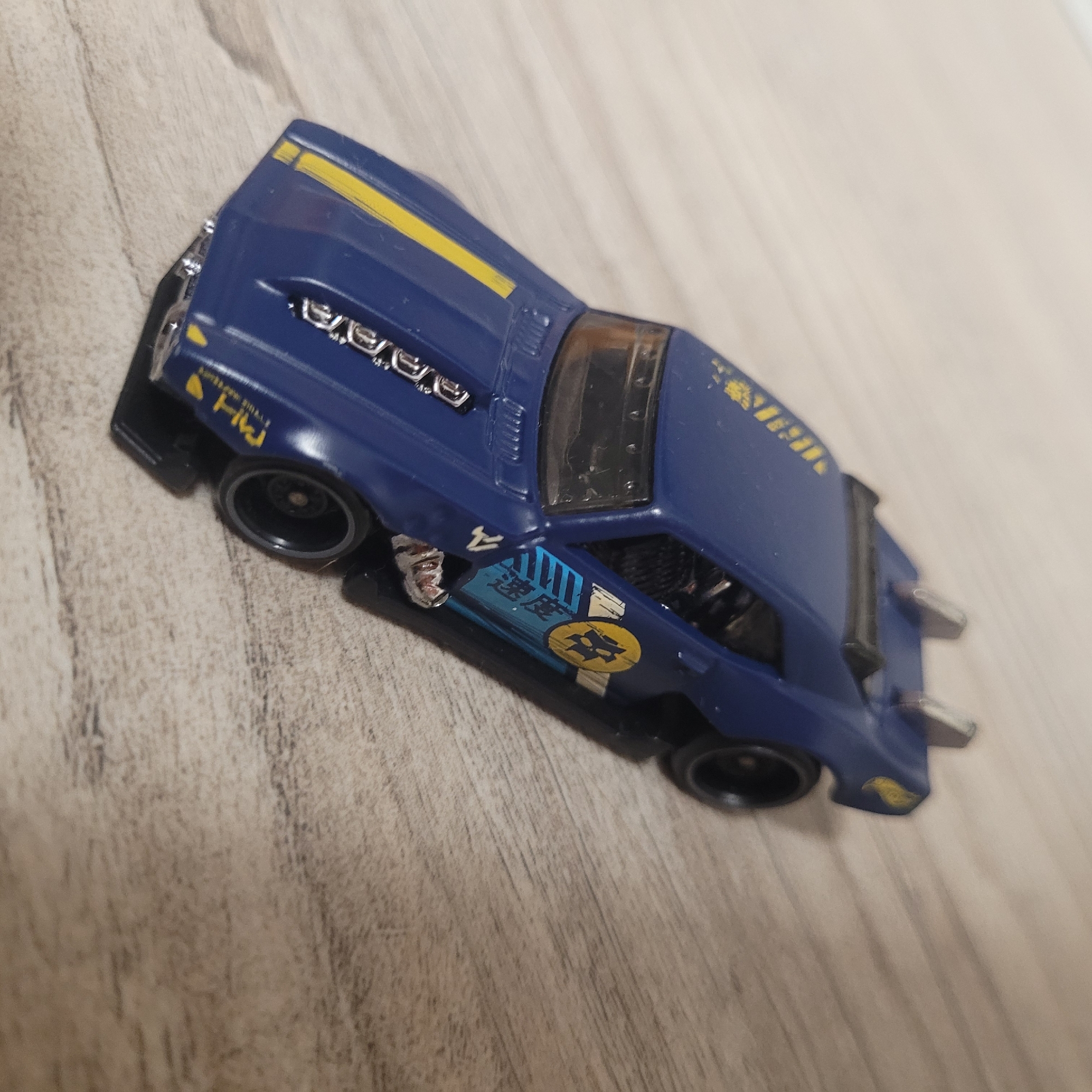 A blue toy car with yellow accents is resting on a wooden surface. The car has several decals, including what appear to be Japanese characters and a circular emblem. The car has visible silver detailing on the hood and what looks like a spoiler or wing on the roof. The background is a light-colored wooden surface with visible grain.