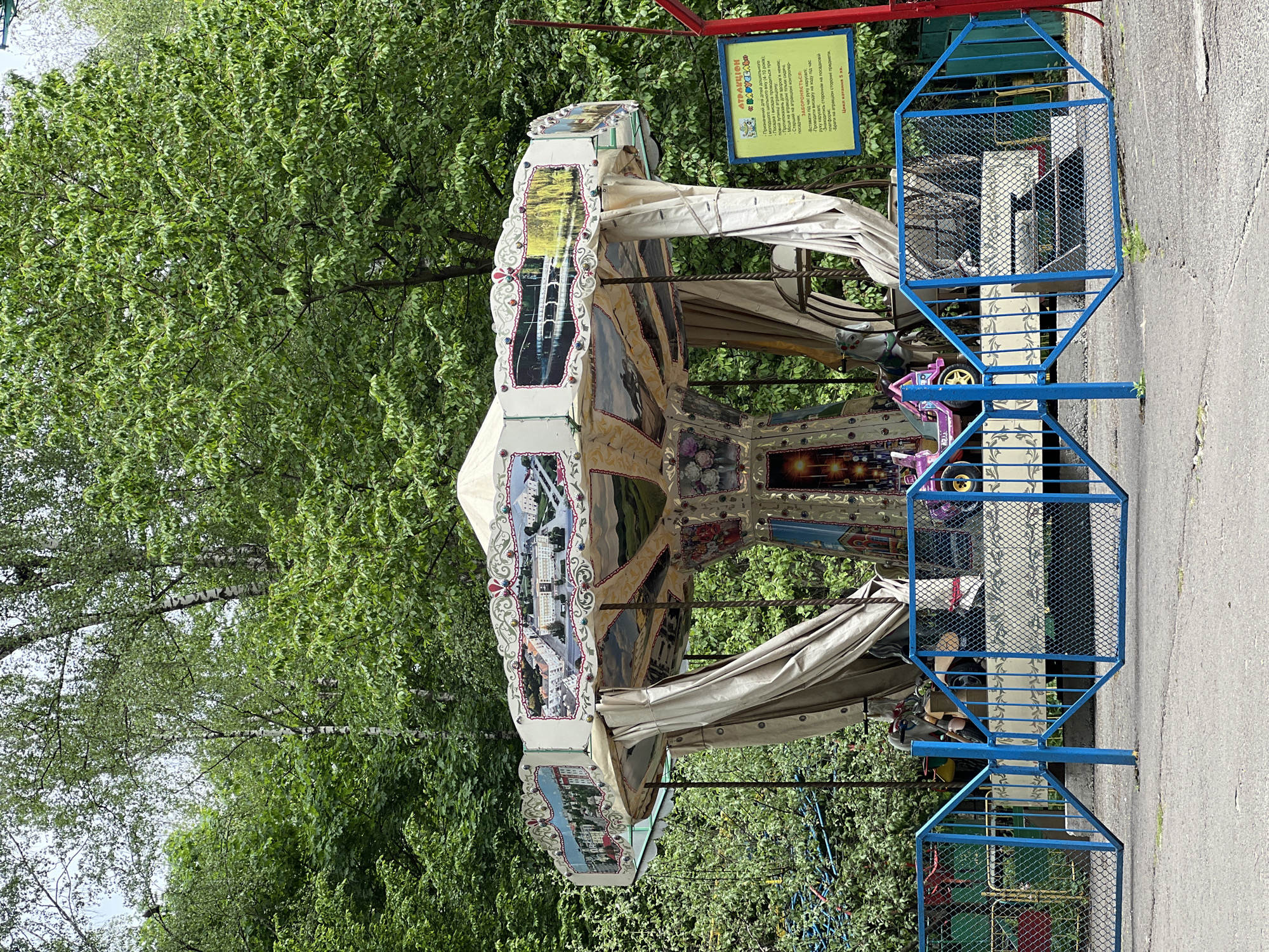 This outdoor shot depicts a carousel at an amusement park in Ternopil, Ukraine. The carousel is decorated with images of various city scenes, including buildings, bridges, and landscapes. The carousel has canvas-like material draped from the sides. Blue metal safety cages surround the ride. A yellow sign with black lettering is mounted behind the carousel, providing instructions and restrictions in Ukrainian. Green trees form the backdrop. The ground appears to be paved. The photo is taken during daylight.
