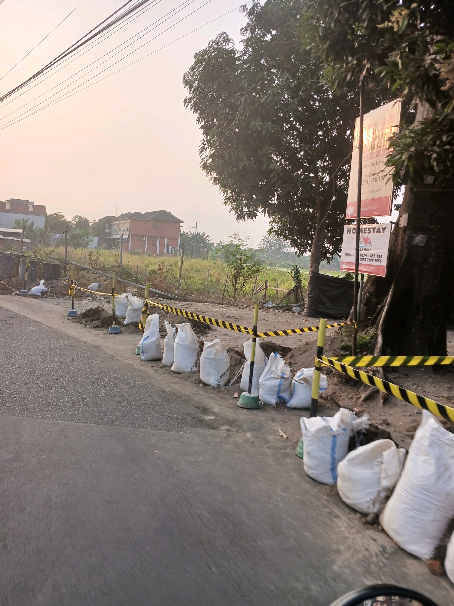 The image depicts a roadside construction scene in Depok, Indonesia, at dawn or dusk, indicated by the soft, warm lighting. A paved road occupies the foreground, with the edge of a vehicle's dashboard and steering wheel visible at the bottom. Alongside the road, a construction area is cordoned off with yellow and black striped tape supported by yellow posts. Several white sandbags are arranged in rows along the edge of the construction site, some appearing partially filled or bulging. Behind the sandbags, piles of dirt are visible, suggesting ongoing excavation or earthwork. In the background, across a grassy field with scattered vegetation, stand two multi-story buildings, one with a reddish-brown facade and prominent white columns. A large tree with dense foliage is situated to the right of the frame, partially obscuring a signpost. The signpost displays two rectangular panels; the top one is largely unreadable due to glare, while the bottom panel, also with some glare, features the word "HOMESTAY" prominently, followed by what appears to be contact information. Power lines stretch across the pale sky overhead. The overall atmosphere is quiet and still, characteristic of early morning or late evening in a semi-urban or developing area.