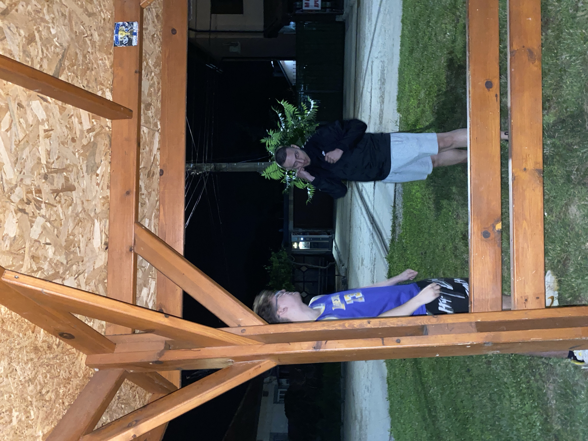 The image shows two young men standing under a wooden canopy at night. The man on the left, wearing a blue basketball jersey and patterned shorts, is partially obscured by a wooden support beam. He is looking to his right. The man on the right, wearing a black jacket and light-colored shorts, is facing the camera and appears to be adjusting something around his neck. He has his fists raised slightly. Behind them, a dark, tree-like shrub is visible, and beyond that, houses and streetlights are dimly illuminated against the night sky. The location appears to be outdoors in a residential area, possibly a yard or a public space. The ground is covered in grass, with a paved area or pathway behind it. The overall impression is of a casual, nighttime gathering.