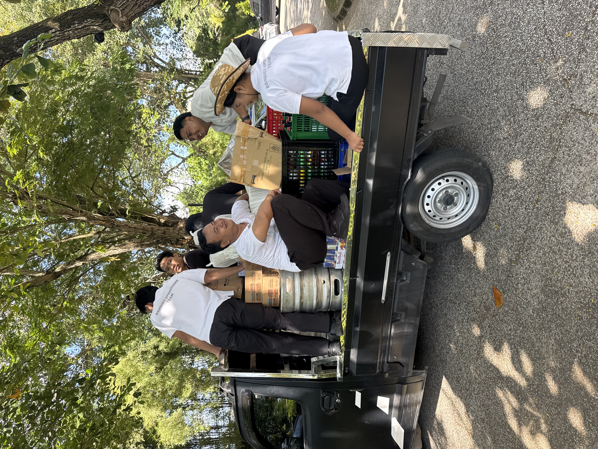 A group of men is captured in an outdoor setting in Kuta Selatan, Indonesia, gathered around and on the back of a black pickup truck, likely engaged in loading or unloading supplies. The image is taken on a bright, sunny day, indicated by clear shadows on the asphalt ground and ample natural light illuminating the scene and the lush green trees overhead.
There are at least five men visible. Most are wearing white t-shirts and dark pants, suggesting a uniform. The text "BANQUET OPERATIONS TEAM" is clearly visible on the back of at least two of the white shirts, indicating their professional role. One man wears a straw hat. Some of the men appear to be actively moving or organizing items, while others are seen resting or casually situated amongst the cargo. One man is lying down on top of some boxes and equipment, appearing relaxed, while another is smiling.
The bed of the black pickup truck is heavily laden with various items. These include numerous cardboard boxes, green and black plastic crates filled with bottles (possibly beverages), several large metal kegs, and cans of Red Bull. One cardboard box is marked with "SANUKI UDON 200G HALAL 調岐うどん," indicating food items. The truck itself is a compact pickup model with a silver diamond-plate trim on the side. The ground is asphalt, and the background is dominated by large, leafy trees, contributing to the outdoor, possibly park-like or service area setting.