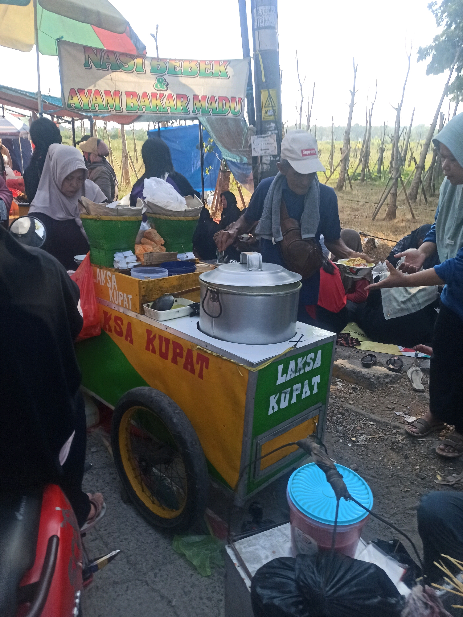 A street food cart is set up in Telagamurni, Indonesia, with a banner advertising "Nasi Bebek & Ayam Bakar Madu" (Duck Rice & Honey Grilled Chicken). A man wearing a white "Supreme" baseball cap and a grey scarf over his shoulders is serving food from a large metal pot. He is holding a bowl of food and interacting with a person whose hands are visible. Several other people, some wearing headscarves, are gathered around the cart. The cart itself is colorful, with yellow and green sections, and displays the words "LAKSA KUPAT" on its side. There are green baskets holding food items, plastic bags, and a blue-lidded container on the cart. In the background, there are trees and vegetation, suggesting an outdoor, possibly semi-rural, setting within the city. The ground is covered in gravel and dirt.