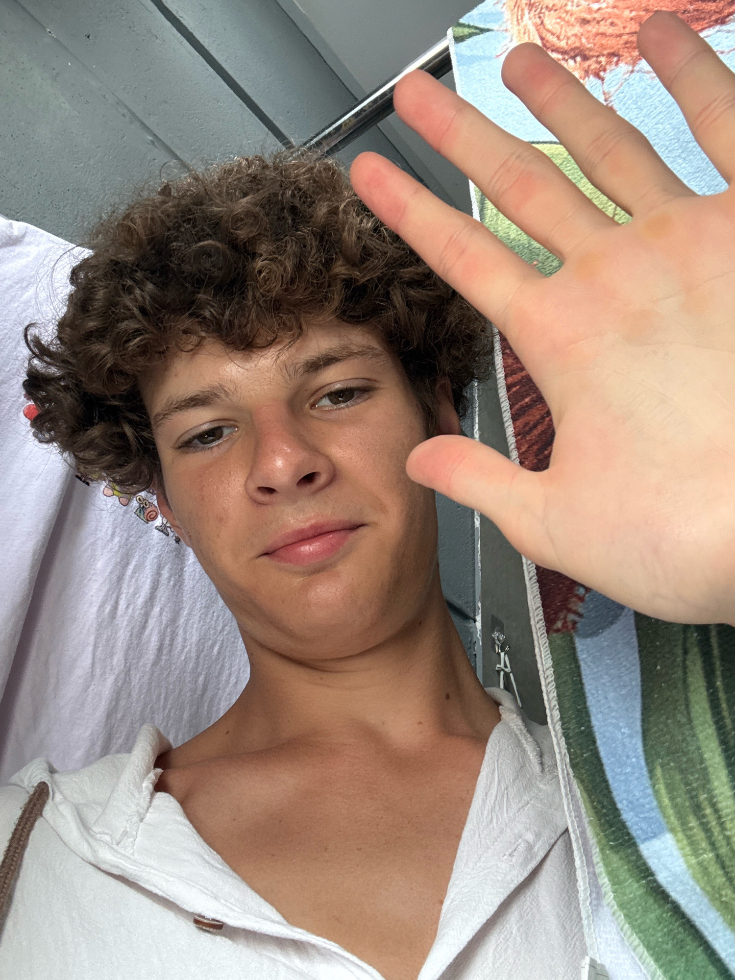 A young male with curly brown hair is seen in a close-up selfie. He is wearing a white hooded shirt. His right hand is raised, palm facing the camera, with fingers slightly spread. The background is out of focus, showing a patterned cloth on the right and a light-colored fabric on the left. The lighting suggests an outdoor setting, possibly during the daytime. The image does not provide any specific visual cues or landmarks that would allow for identification of the location within Nha Trang, Vietnam.