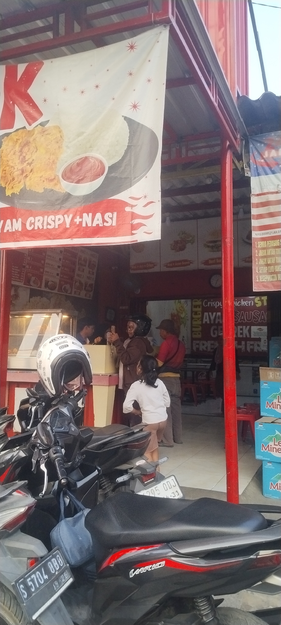 The image shows a street-level view of a food stall in Bedah Lawak, Indonesia. The stall appears to be selling fried chicken and rice. A large banner advertising "AYAM CRISPY + NASI" is prominently displayed. In the foreground, several motorcycles are parked, one with a white helmet on its handlebars. Several people are visible interacting around the stall, including a woman in a brown jacket and headscarf, a younger girl in a white shirt, and a man in a red shirt. A display case filled with food items is visible inside the stall. Stacks of bottled water, likely for sale, are positioned to the right of the stall. The overall scene suggests a busy commercial area typical of a local market or street food vendor.
