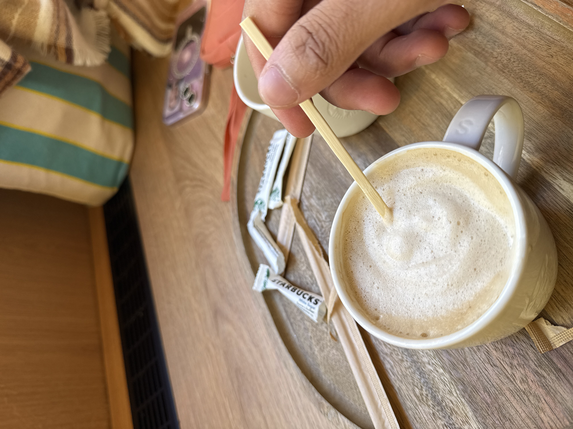 A close-up, high-angle shot captures a person stirring a frothy beverage in a white mug set on a light wooden tray or table. A hand is visible from the wrist down, holding a light wooden or bamboo stirrer deep into the beige-colored, foam-topped drink, likely a latte or cappuccino. The mug has a light grey handle, featuring a subtle "S" embossed on it.
Scattered on the wooden surface are several sugar packets, including white ones clearly branded "STARBUCKS White Sugar," and at least one brown sugar packet. A small, empty white ceramic cup is also visible near the stirrer. To the left of the mug, partially in focus, lies a striped cushion or pillow with wide beige, light blue, and thin yellow stripes. Above it, a blurred striped blanket or throw with fringed edges adds to the cozy atmosphere. A smartphone with a distinct purple case and prominent camera lenses is partially visible near the cushion. Along the bottom left edge, a dark, gridded vent or grate suggests an integrated furniture feature or floor detail.
The scene depicts a relaxed moment of enjoying a drink, likely at a café, home, or office in Paris, France, given the contextual information. The Starbucks branding indicates the origin of the beverage or accompanying items. The lighting suggests daytime.