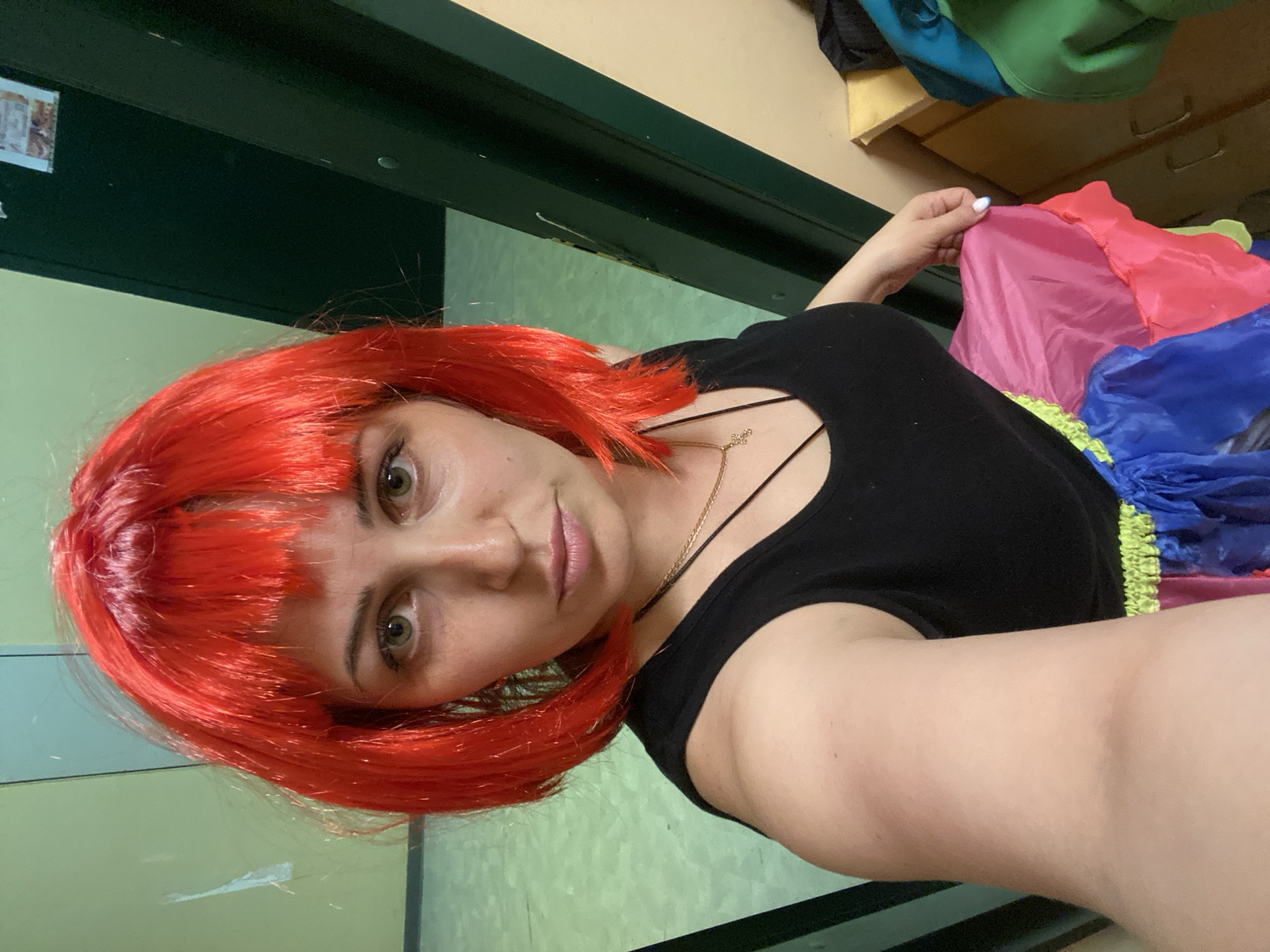 A selfie captures a woman indoors in Марковский, Russia, looking directly into the camera with a neutral expression. She is wearing a vibrant red wig with bangs, a black top with thin straps, and a delicate gold-colored necklace. Her eyes are green.
The setting appears to be an interior space, possibly a functional room or backstage area, with a dark green door frame or panel running along the left side and a light green wall in the background. A small, blurry rectangular object, possibly a poster or sign, is barely discernible on the upper left wall.
On the right side of the frame, the woman holds a brightly colored, multi-textured fabric, displaying shades of pink, blue, and a ruffled yellow-green trim, which may be part of a costume or prop. Above this, wooden drawers or shelves are visible, along with blue and green bags or items of clothing. The lighting suggests an indoor environment, and the woman's pose indicates she is actively taking the photograph. Her visible hand shows light-colored nail polish.