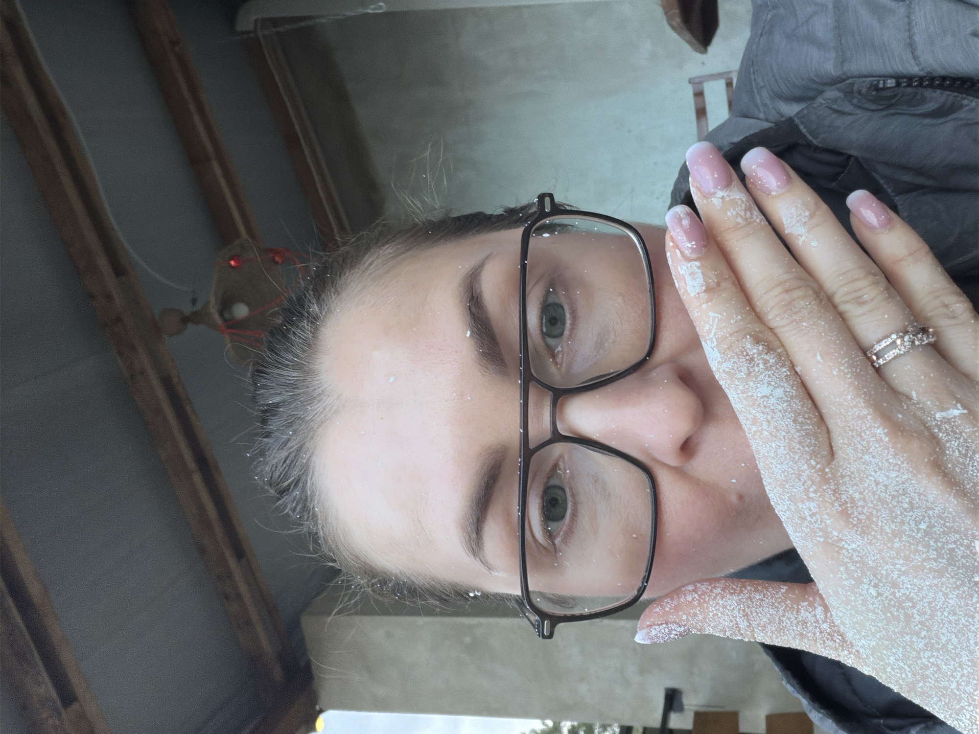 A close-up, selfie-style image shows a woman's face and hand covered in a white, powdery substance, potentially snow or flour. The woman has fair skin, green eyes, and is wearing black-framed glasses. Her nails are manicured with a pink and white design, and she is wearing a ring on her ring finger. She is wearing a dark-colored jacket. The background is a blurry mix of natural and built elements, including what appears to be an angled roof structure. The specific location in Dubovaya, Russia cannot be determined from the image.