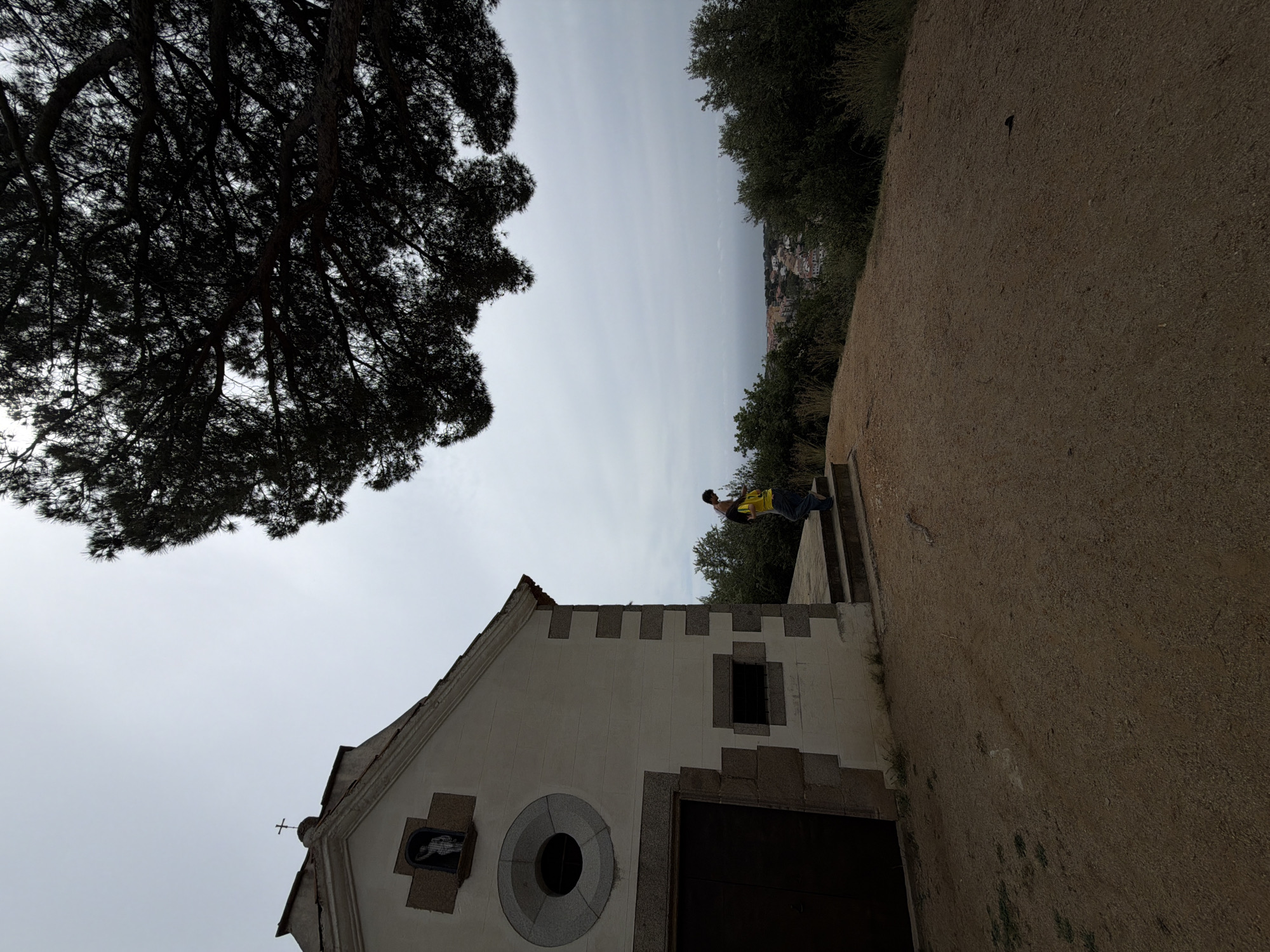 A low-angle shot shows a person walking down stone steps past a white building with a dark-colored door, stone accents, and a cross on the roof, with a large tree on the left. The building appears to be at the top of a hill, with a view of a city in the distance. To the right of the building is a path with dirt or sand. The sky is overcast. The location is the chapel of the Sant Andreu de Llavaneres cemetery in the city of Sant Andreu de Llavaneres, Spain.