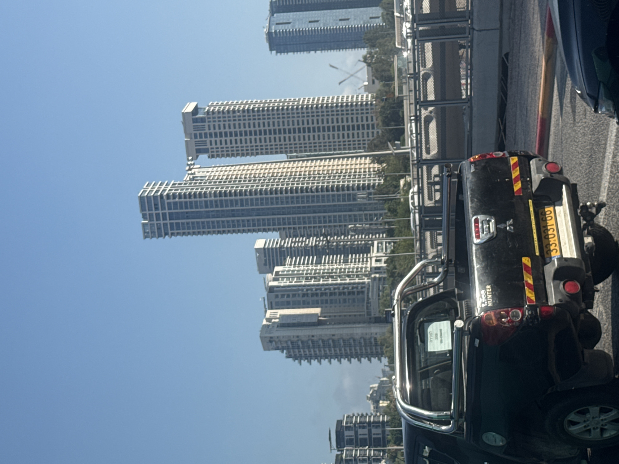 The image shows a black Mitsubishi L200 Hunter pickup truck parked on a road in Tel Aviv, Israel. The truck has a chrome roll bar and a yellow license plate with the number 33:031:66. In the background, there are several tall buildings, including skyscrapers, and a clear blue sky. The scene appears to be a sunny day, and there are other vehicles and trees visible in the distance. A white sign with Hebrew text is attached to the rear window of the truck.