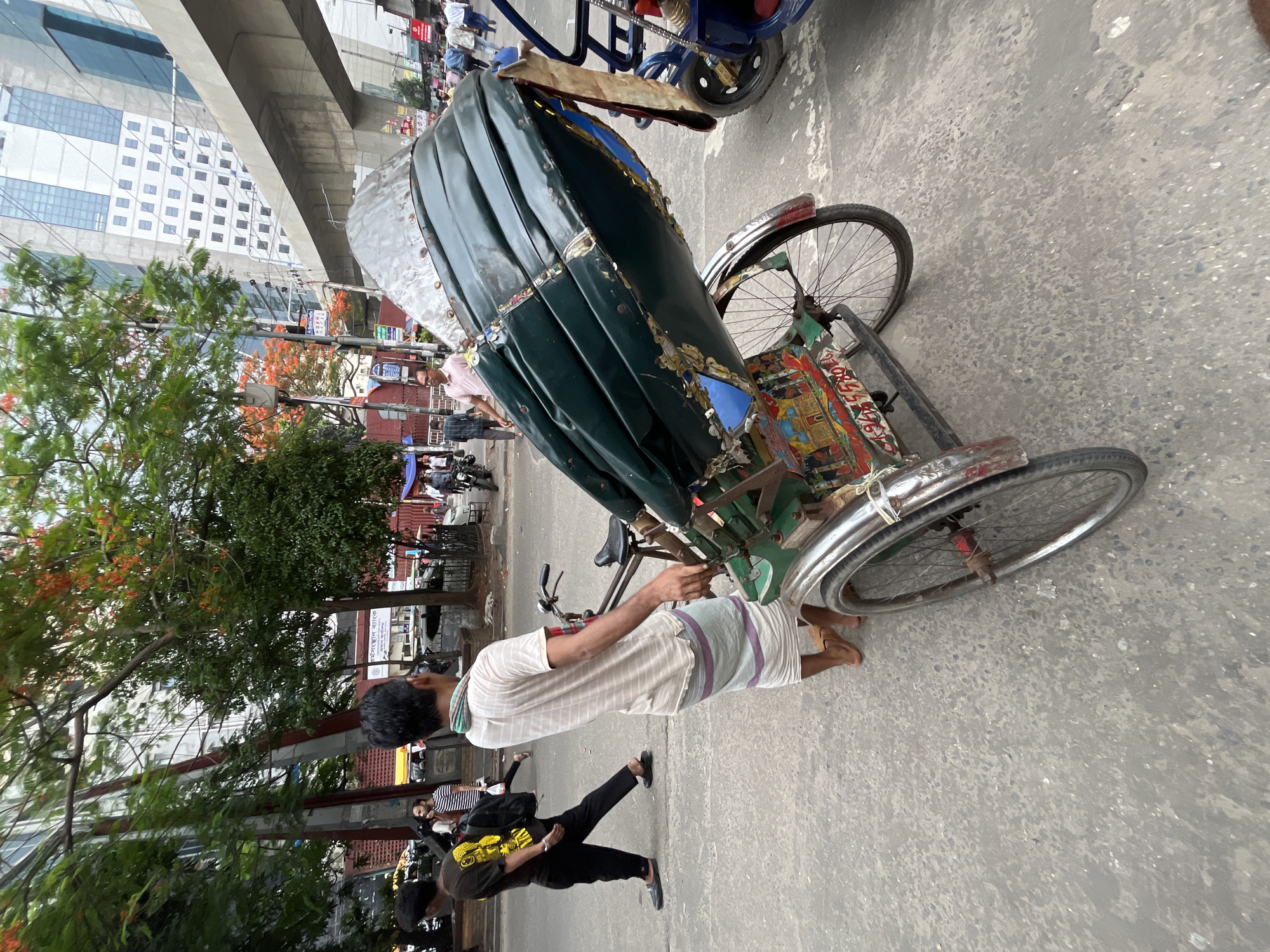 In a street scene in Dhaka, Bangladesh, a rickshaw puller in a striped shirt and lungi stands next to his rickshaw. The rickshaw has a green canopy. Two people are walking in the background. A building and a roadway are behind them.