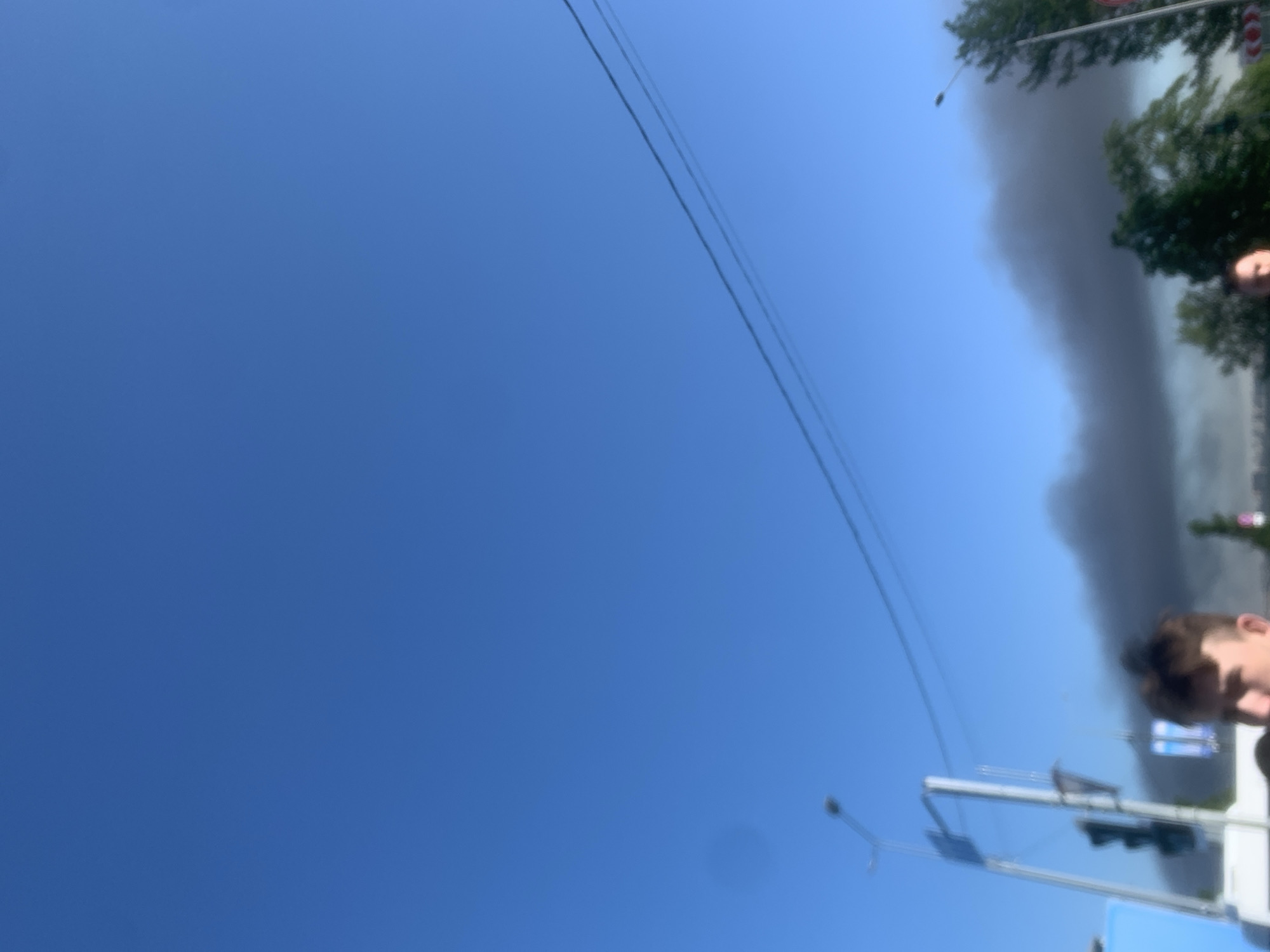 The image, taken in Shushary, Russia, prominently features a vast expanse of clear, bright blue sky dominating the upper and left portions of the frame. On the right side, a substantial, dark grey to black plume of smoke rises vertically, contrasting sharply with the azure sky and suggesting a fire or significant emission event.
Partially visible on the far right, intermingled with the smoke, are clusters of green tree foliage. Two thin, dark overhead wires or power lines traverse diagonally across the upper-middle section of the sky. Towards the lower right, there are blurry indications of street infrastructure, including parts of a metal pole that could be a streetlight or traffic signal. A very blurry human head, seen in profile and partially cropped, is present in the extreme lower-right corner, seemingly looking towards the scene unfolding.
The photograph was taken during daytime under fair weather conditions, with the sun illuminating the sky. The overall impression is one of an incident (likely a fire) occurring in an urban or suburban setting, observed by an individual. No visible text is present in the image.