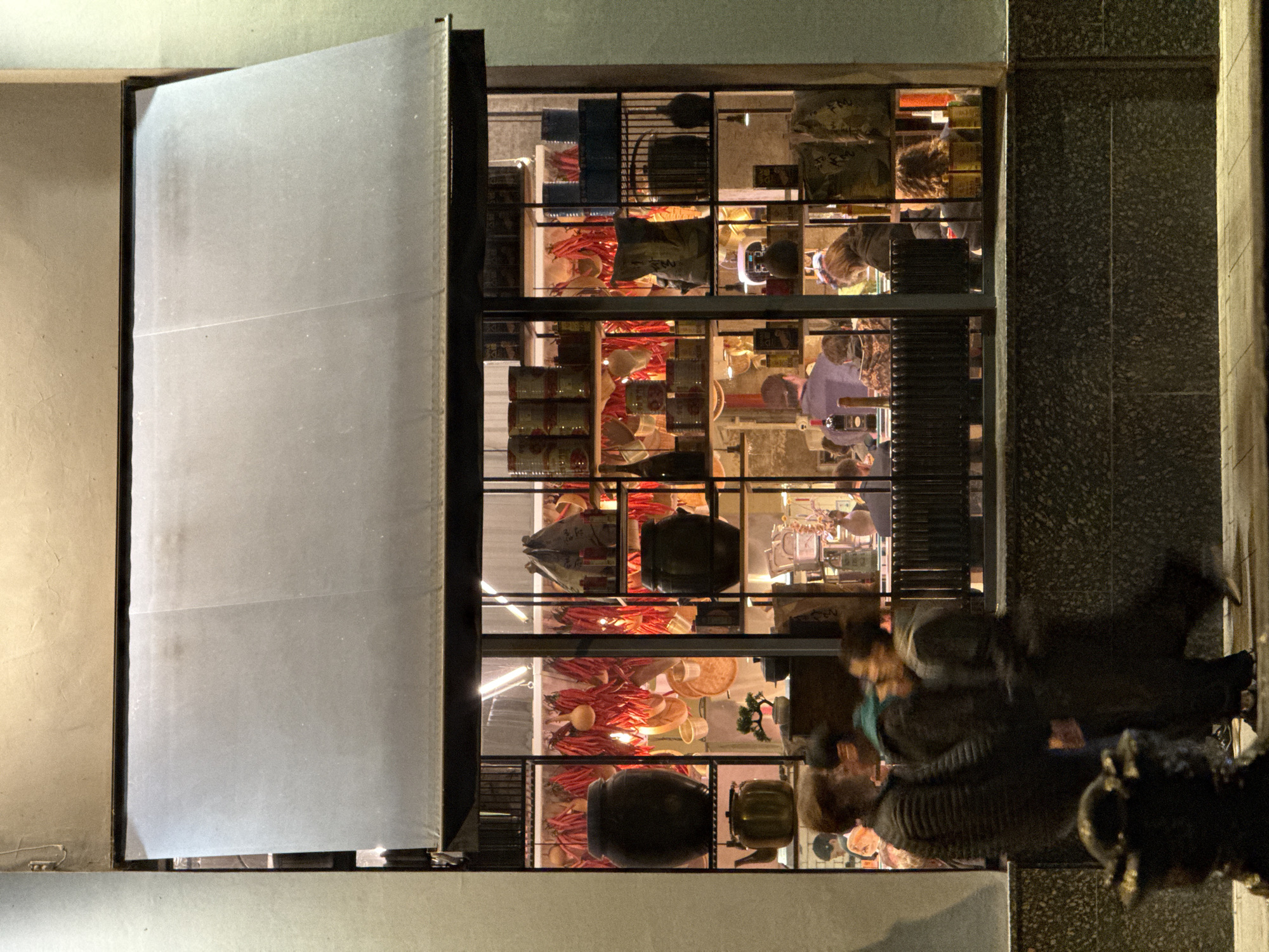 An eye-level, rotated, medium shot shows a commercial display window and open door or panel of a building in Moscow, Russia. The window is illuminated from within, showing shelves stocked with red peppers, canned goods, and other miscellaneous items. People are inside the shop. A few blurry people pass by on the street outside the window. An open panel or door is on the left of the window. The exterior wall is made of concrete-looking material.