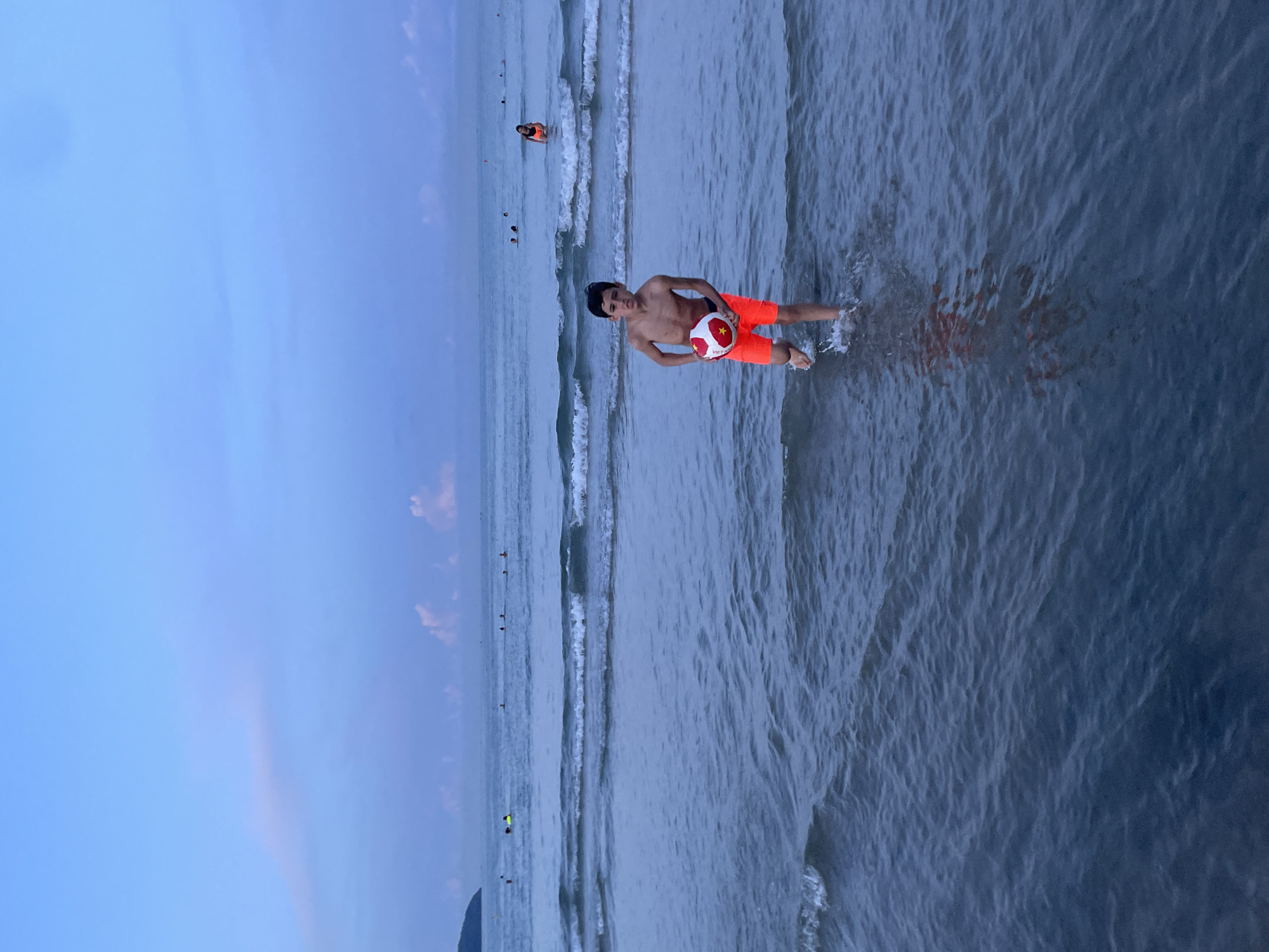 A young boy with dark hair stands in the shallow water of the ocean at My Khe Beach in Da Nang, Vietnam. He is shirtless and wearing bright orange swim trunks, and he holds a red and white striped ball. The water is calm with gentle waves, and the sky is a soft blue with hints of pink from the setting sun. Other people are visible in the distance, swimming and wading in the sea. The overall scene conveys a sense of peacefulness and a beautiful evening at the beach.