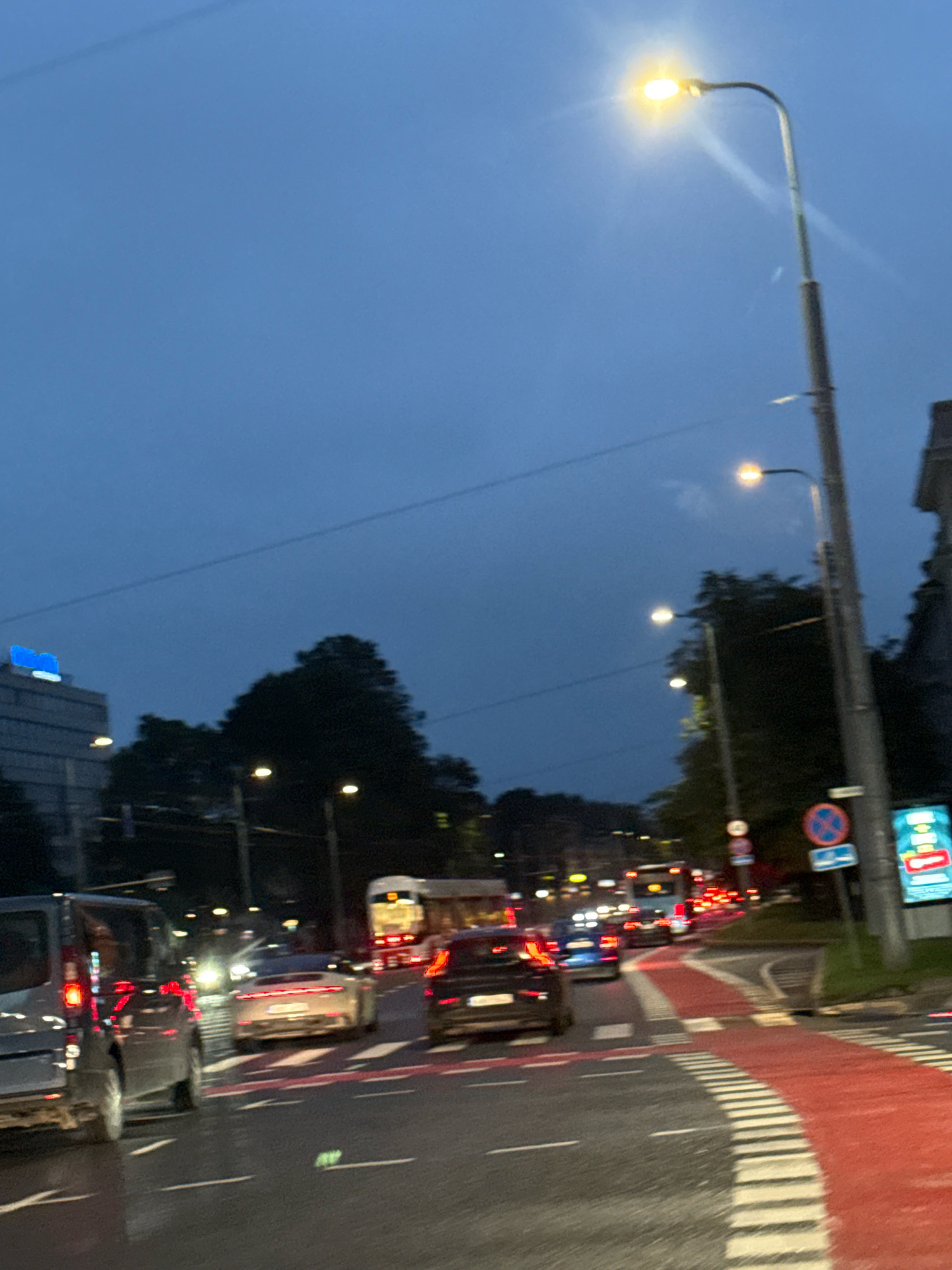 The image shows a street scene in Tallinn, Estonia, at dusk or night. A street light illuminates the dark blue sky. Traffic is visible, including cars, a bus, and a van. The road has distinct red and white markings, indicating a bike lane or pedestrian crossing. Buildings and trees are present in the background, with one building displaying a bright blue sign. The overall impression is of a city street during evening hours with moderate traffic.