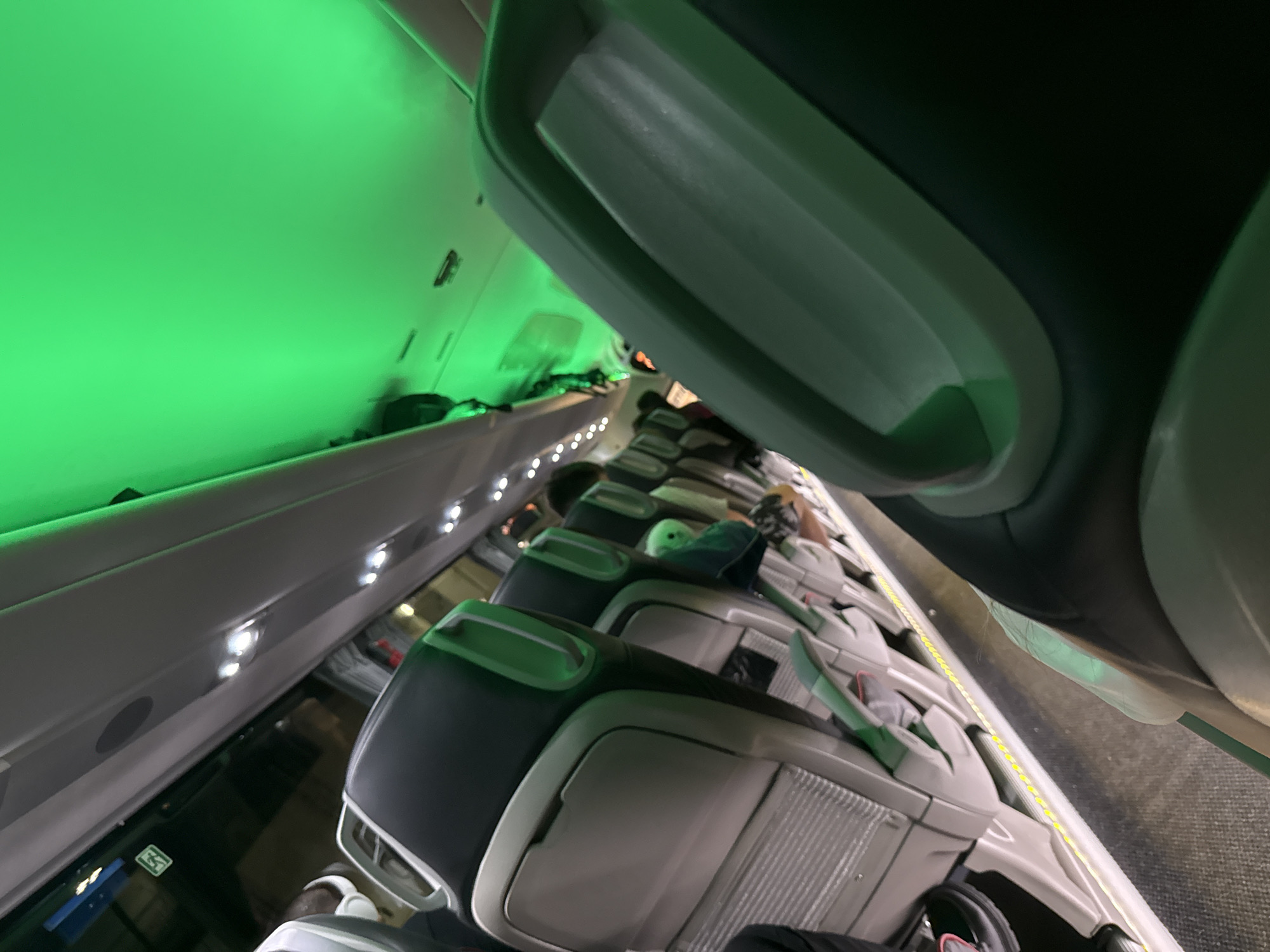 The image shows the interior of an aircraft cabin, viewed from a seat. The cabin is lit with green mood lighting. Rows of seats extend into the distance, some occupied by passengers. Overhead storage bins are visible above the seats. There are no people, objects, actions, interactions, or environmental details to place this image in Varna, Bulgaria.