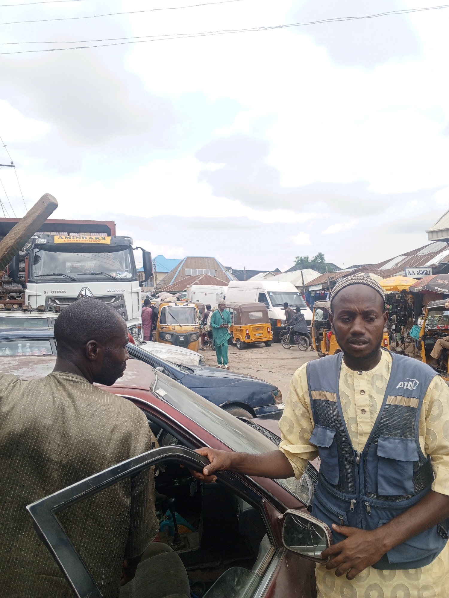 The image captures a busy scene in an urban environment, presumably a market or transport hub in the city of Unknown, Unknown. A man wearing a kufi cap and a navy blue vest with an "ATN" logo stands next to a maroon car with an open door and a broken side mirror. Another man, his back to the camera, is near the open car door.
In the background, there's a truck with "AMINBAKS TRANSPORT" printed on it. Several "Keke Napep" tricycles are visible, as well as other cars, motorcycles, and pedestrians. There is a building with the name "A.A. ACHIDA" visible. The sky is overcast.