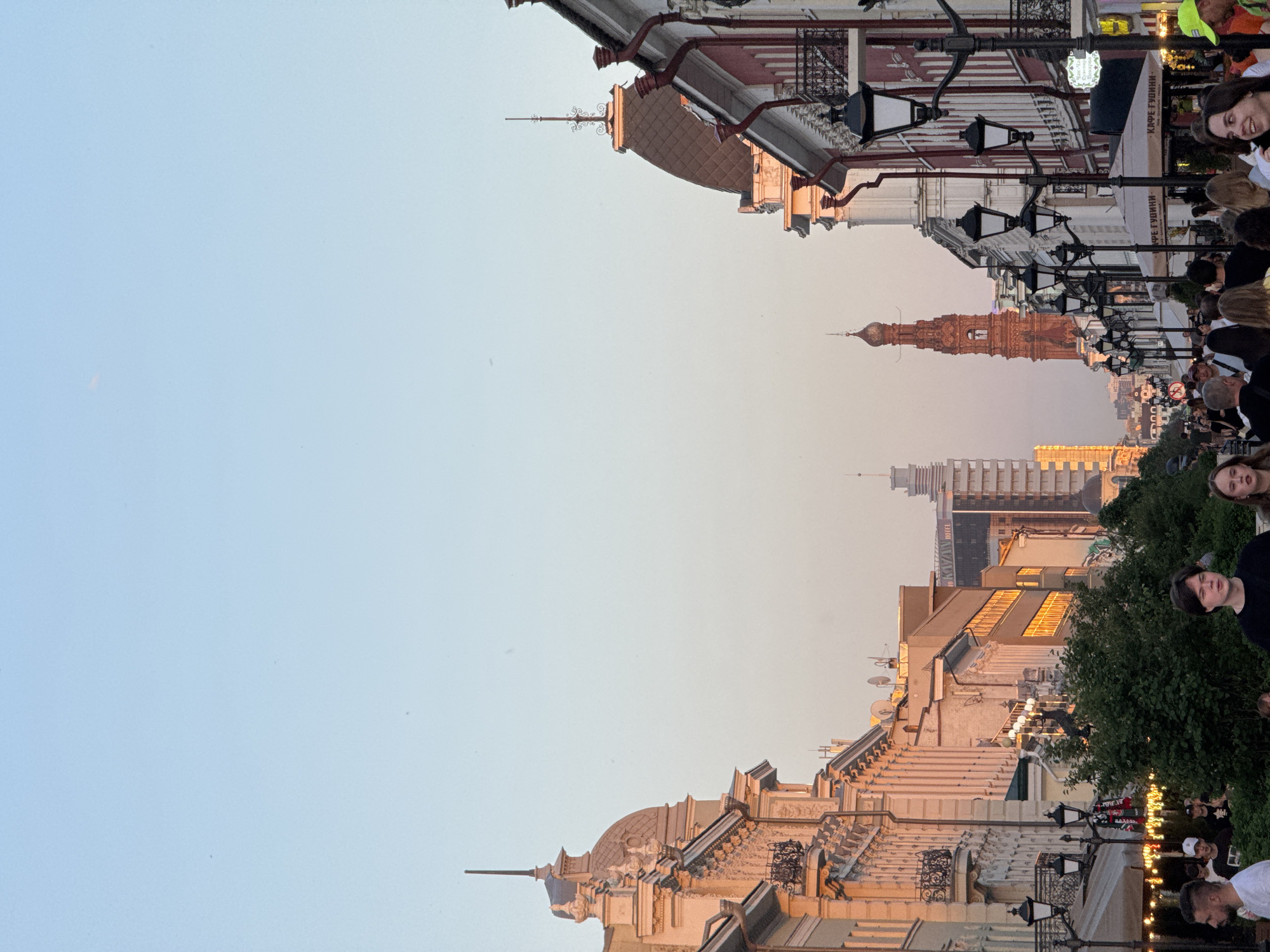 This outdoor shot depicts a street view in Kazan, Russia. The architectural details on the buildings along the street are visible. People are present, some partially obscured on the street. Street lights line the path between the buildings. In the distance, a multi-tiered tower, likely a cathedral, rises above other structures. A taller, modern building is also visible in the background. The sky is a pale blue.