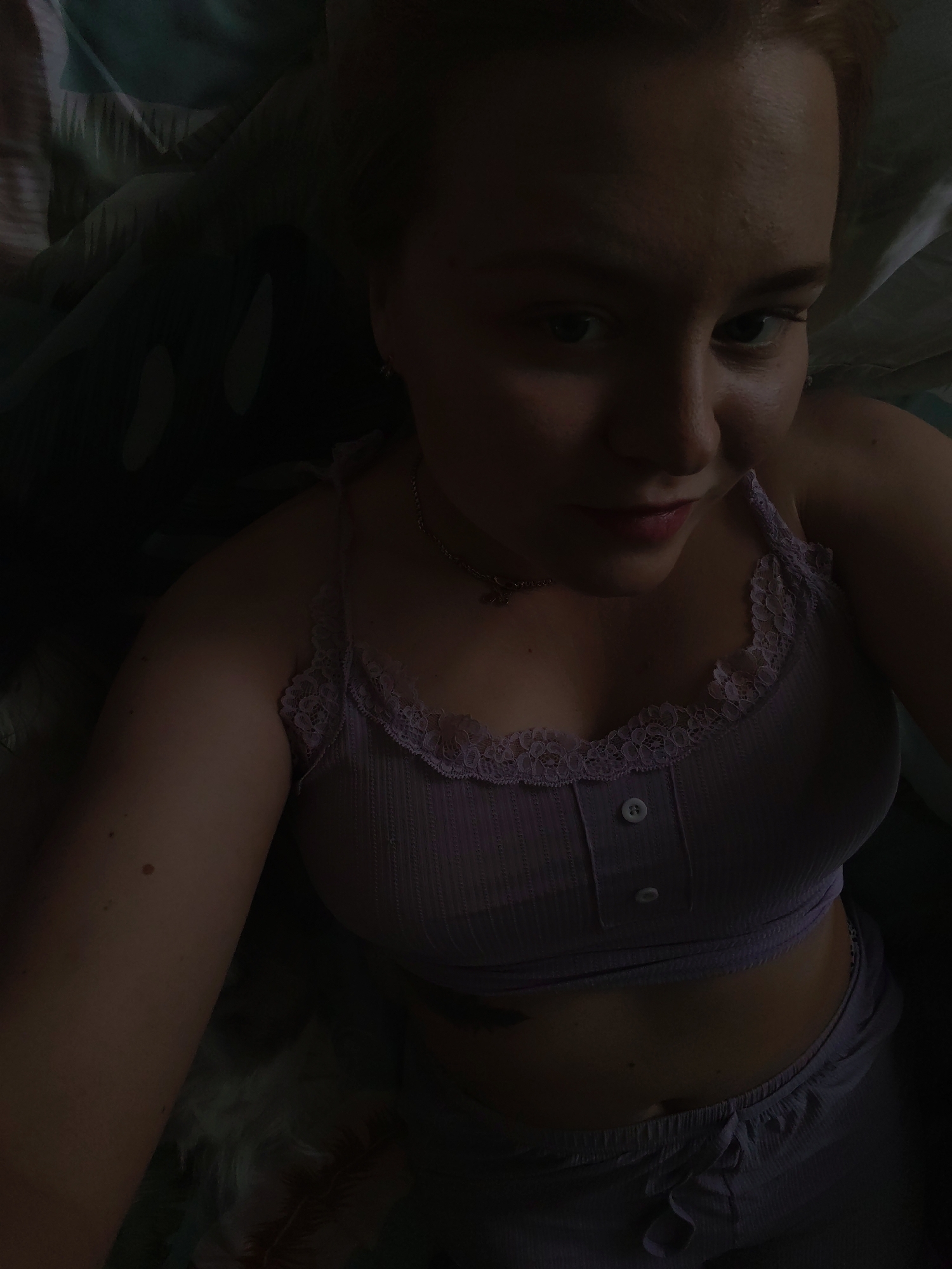 A woman with fair skin and reddish-blonde hair is pictured from a high angle. She wears a light purple crop top with lace trim and buttons, paired with matching drawstring shorts. A silver necklace is visible around her neck. The background includes bedding with a dark, patterned design. The lighting is dim. The location is not discernible.