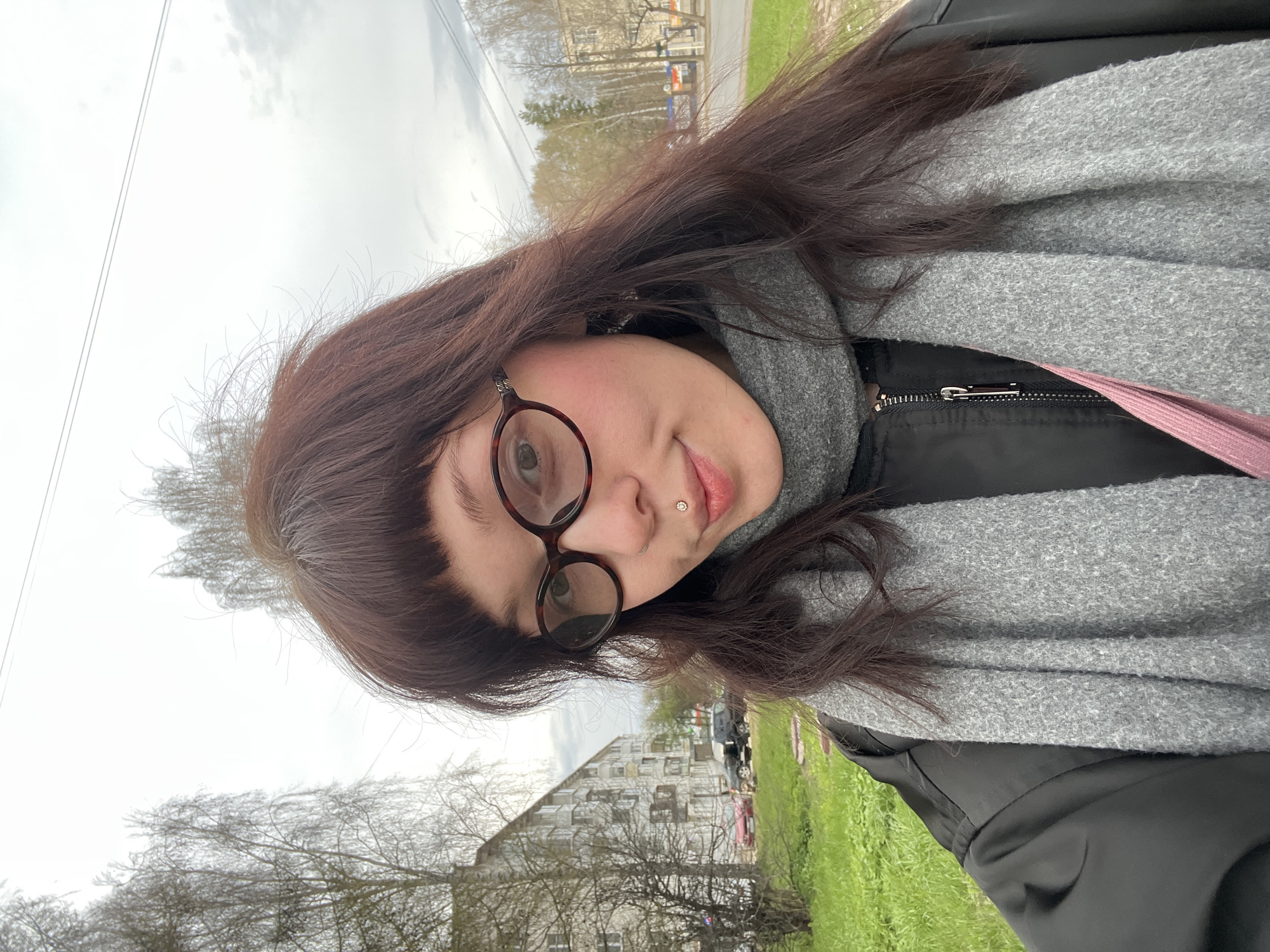 A woman with dark brown hair, round glasses, and a lip piercing is posing outdoors. She is wearing a black jacket and a gray scarf. The background includes a residential building, trees with budding leaves, and a patch of green grass. Based on the location identification request, this image was likely taken in the city of Chaikovsky, Russia.