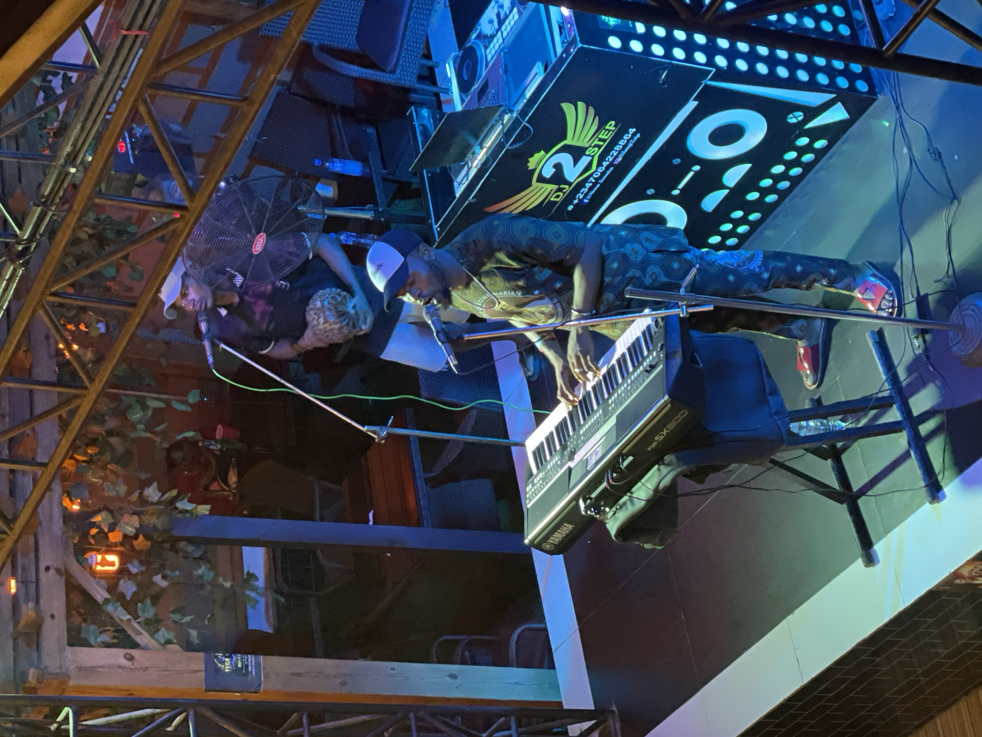 A man is playing a Yamaha SX600 keyboard on a stage. Another man stands to his left, holding a maraca. In the background is DJ equipment with "2 Step DJ" printed on it, including a phone number with a Nigerian country code (+234). A fan is positioned behind the man with the maraca. The stage is decorated with scaffolding and artificial foliage. The scene takes place in Obosi, Nigeria.