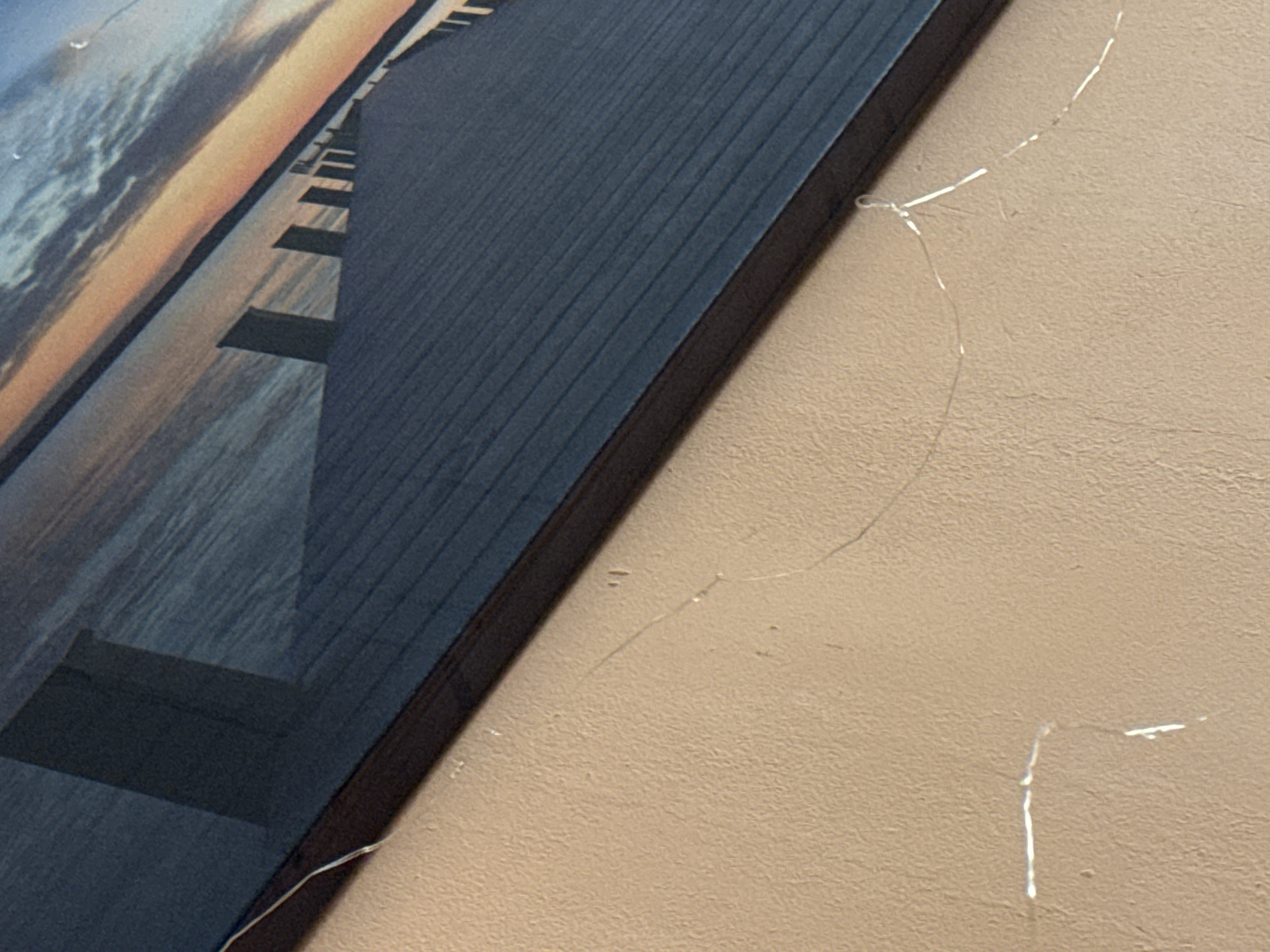 A close-up shows a portion of a framed picture hanging on a beige textured wall. The picture features a scene of water and sky at sunset, with a pier or dock extending into the water. The frame has a dark brown edge. A thin, translucent strand is visible, running from the frame and across the wall, appearing to be detached in several places. Some areas on the wall appear damaged.