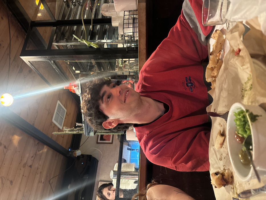 A young man in a red sweater sits at a table with food. He's smiling towards the camera. His sweater has a small logo on the chest. A partially eaten meal with pita bread and a bowl containing lettuce and olives sits on the table. The background shows wooden walls, a mirrored structure, and a reflection of a woman in the mirror, looking at the camera. A bright overhead light shines in the upper left corner.