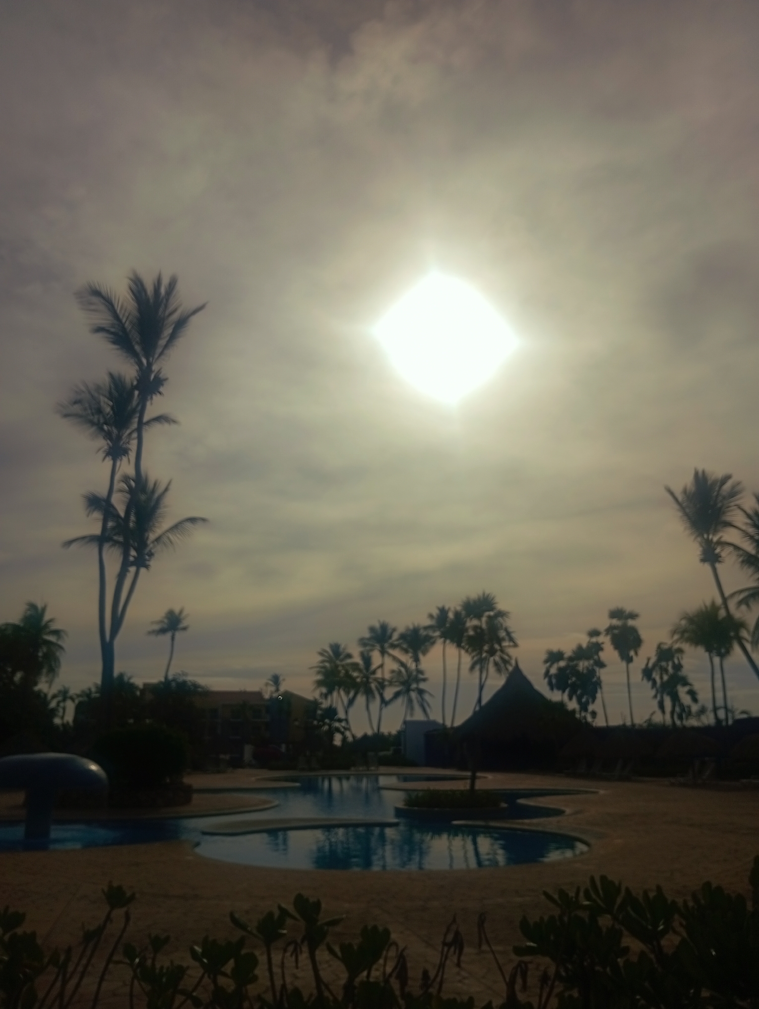 The image depicts a resort scene in Playa El Agua, Venezuela, under a cloudy sky with the sun obscured. A swimming pool complex occupies the midground, bordered by palm trees. A thatched-roof structure and sun loungers are visible adjacent to the pool. In the foreground, vegetation grows along the ground. In the far background, a building is partially visible.