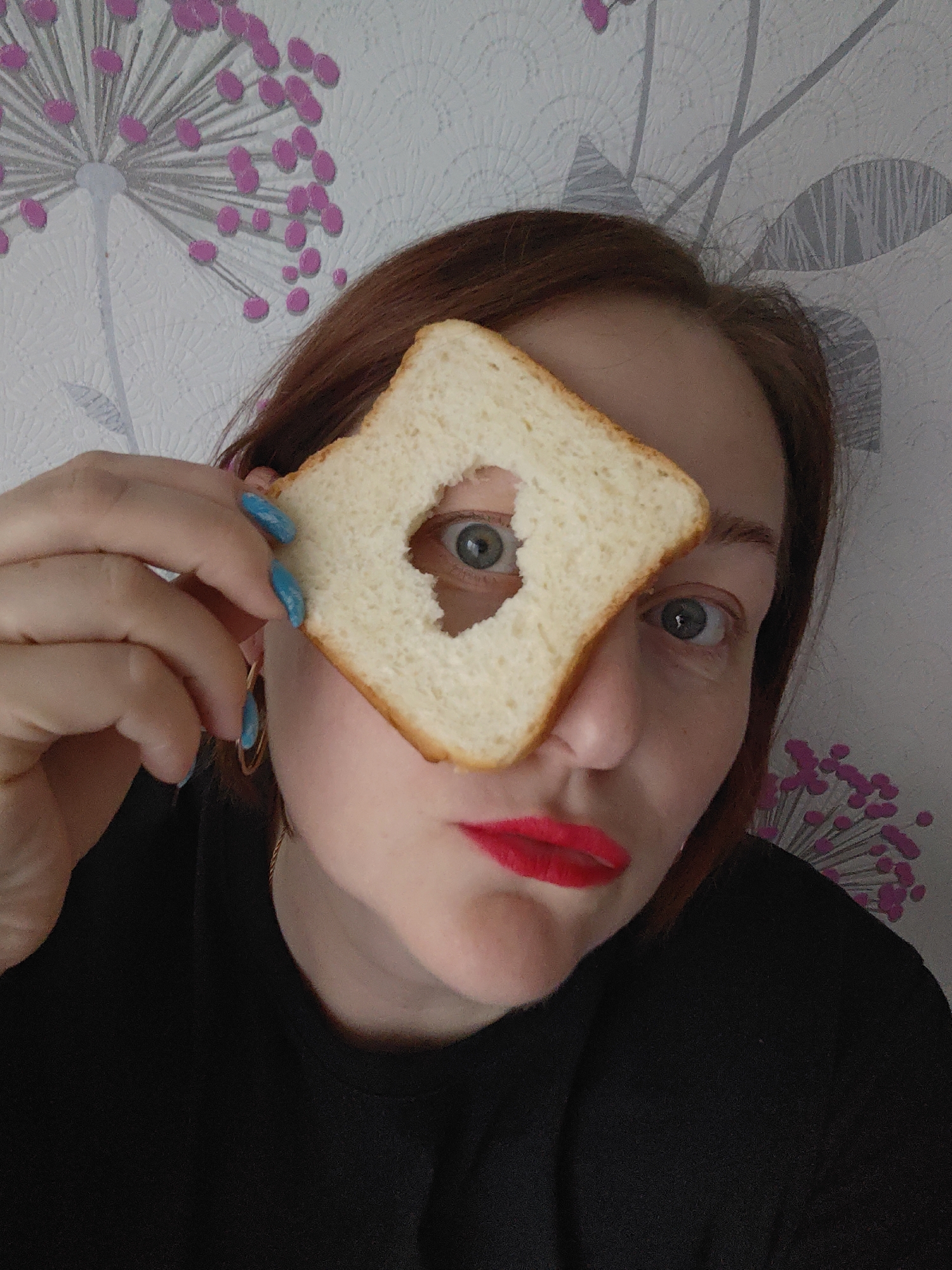 A woman with auburn hair and red lipstick is holding a slice of white bread with a heart-shaped hole cut out of it in front of her left eye. She is wearing a black shirt and gold hoop earrings. Her fingernails are painted turquoise blue. The background is a white wall with a pattern of stylized pink flowers. The photo was taken indoors, most likely in Markovskii, Russia.