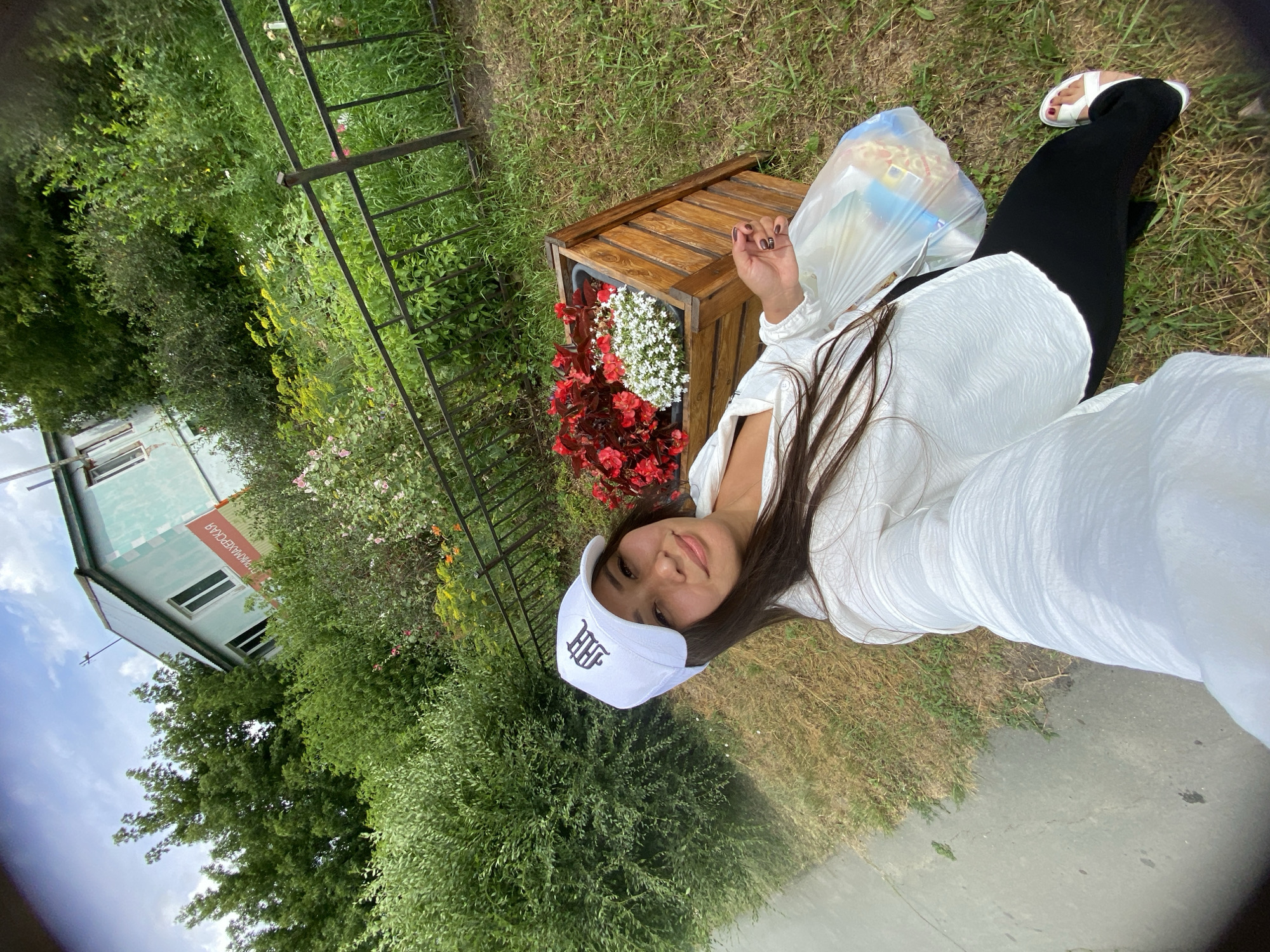 A woman wearing a white baseball cap and a white blouse is taking a selfie outdoors. She is holding a plastic bag in her right hand. Behind her, there is a wooden planter box filled with red and white flowers, a metal fence, and lush green vegetation. In the background, a light blue building with a green roof is visible, with a sign on its side that appears to be in Russian text. The woman is standing on grass next to a paved walkway. The scene is set in a natural or park-like environment within the city of Akyar, Russia.