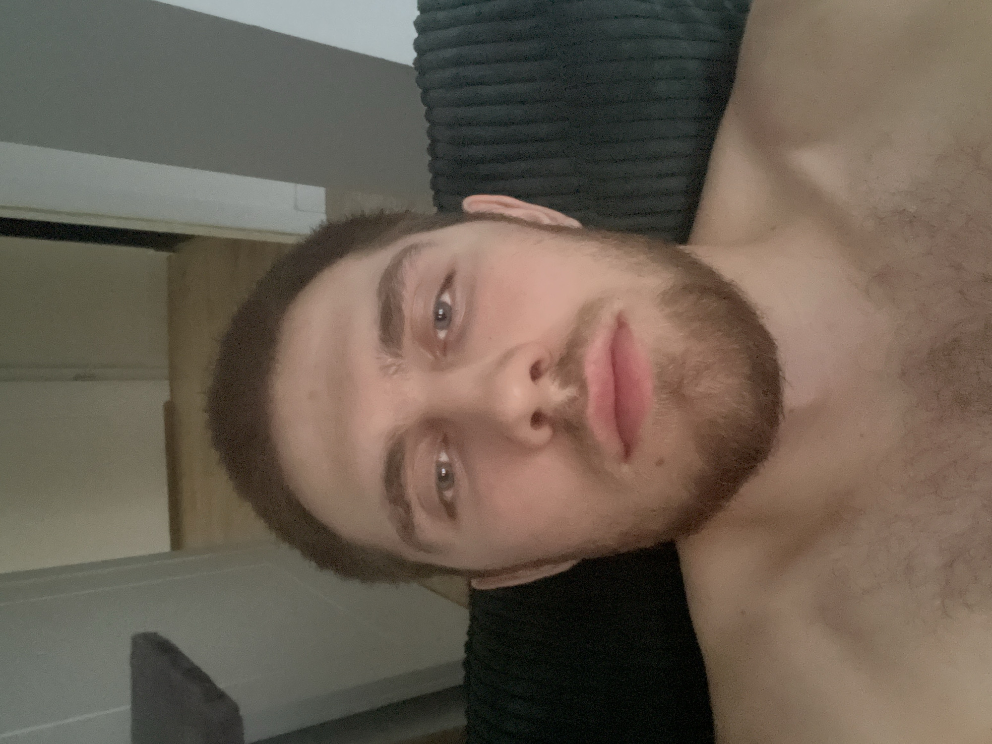 The image is a close-up, selfie-style photograph taken from the chest up of a young adult male with fair skin, brown hair, and a light beard. He is looking directly at the camera. The background is out of focus, but appears to be an indoor setting with a doorway and wooden flooring visible. The man is shirtless, and his skin shows some minor blemishes. The lighting is dim, creating soft shadows on his face. The image does not contain any information about the city of Pruszcz Gdański, Poland.