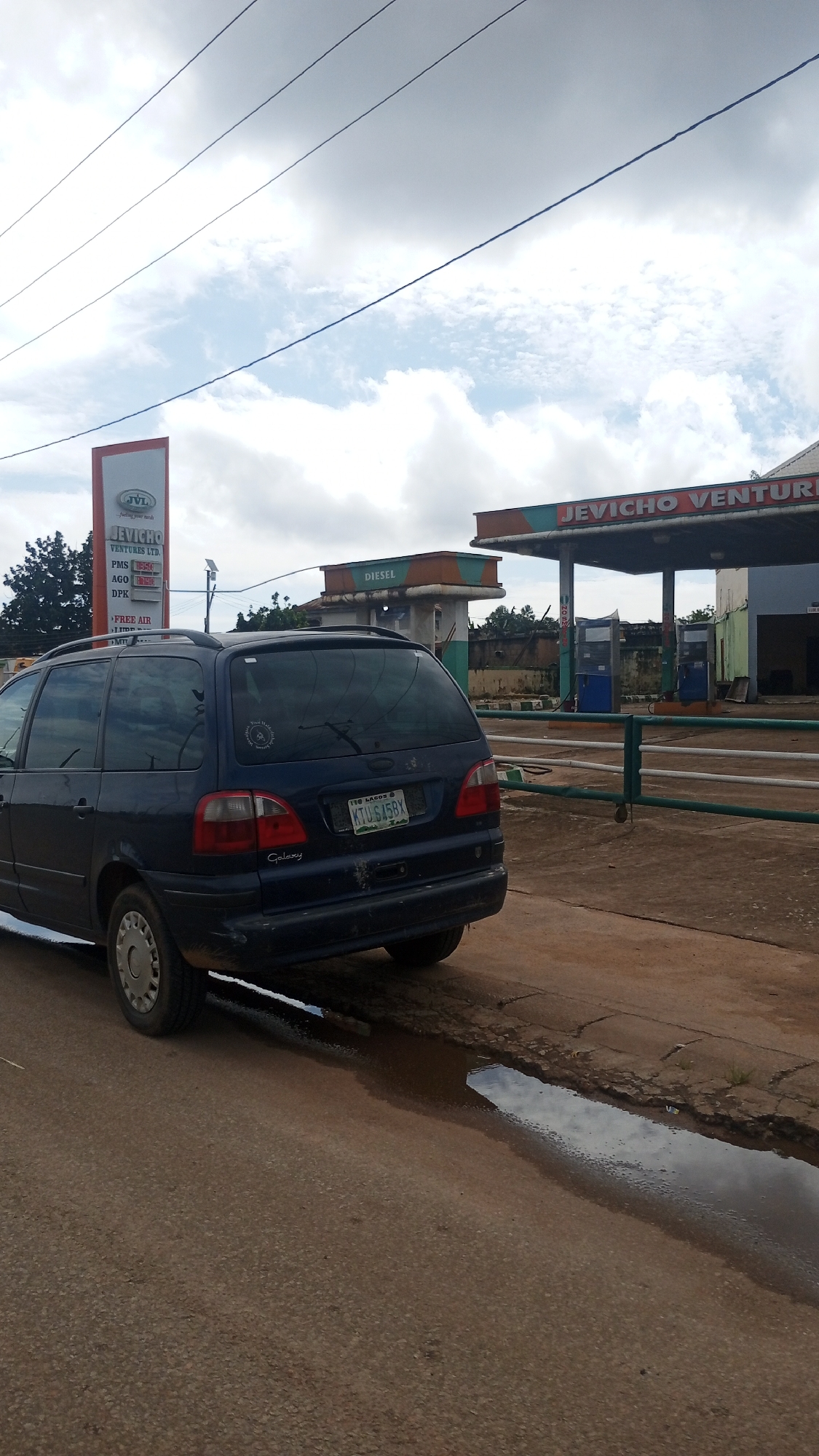 The image depicts a dark blue minivan parked on a dirt road in front of a gas station. The gas station has a prominent sign reading "JEVICHO VENTURES LTD" and "DIESEL". There are several fuel pumps visible under the station's canopy. The minivan has a license plate that reads "KTU 645BX". The road is uneven with a large puddle of water on the right side. The sky is cloudy. The scene appears to be set in Bukuru, Nigeria, as indicated by the mention of this city in the prompt. There are no people or discernable actions in the image. The overall impression is one of a typical roadside scene at a fuel station in a Nigerian city.