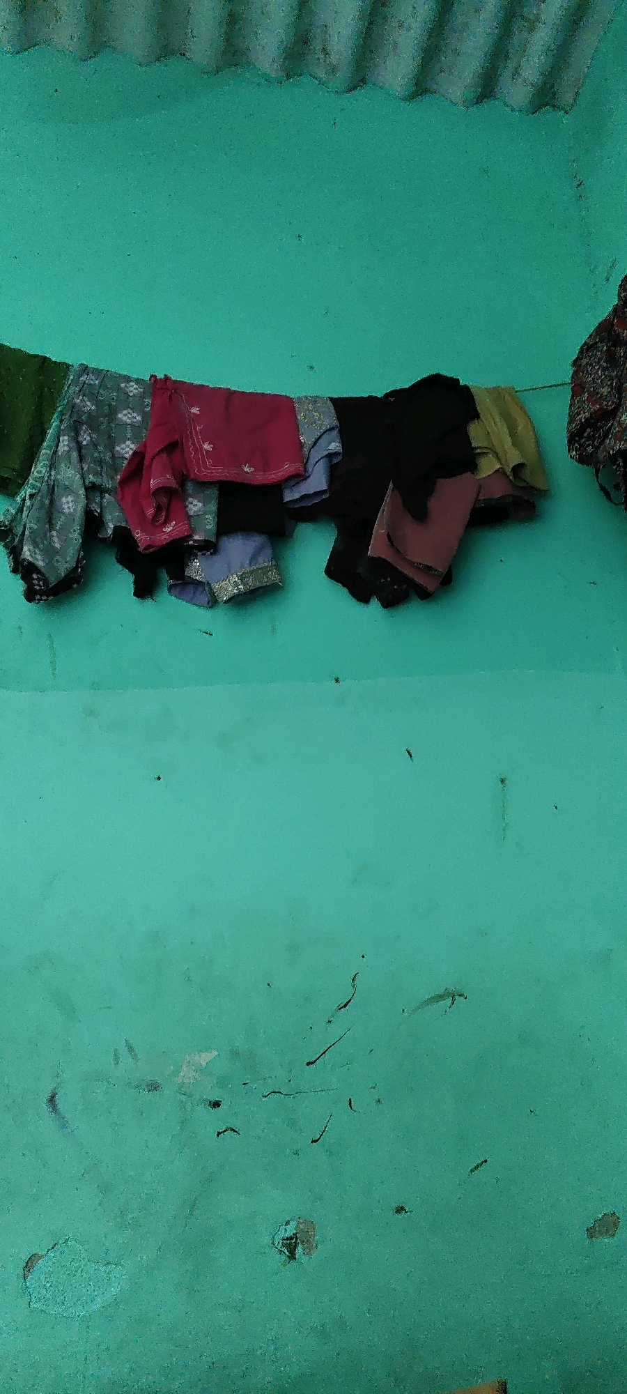 A clothesline is strung horizontally across a weathered, turquoise-painted wall. Various garments, including what appears to be a green dupatta, a printed grey and white fabric, a maroon tunic with intricate embroidery, a black garment, a light green fabric, and a pinkish-brown garment are draped over the line. Above the clothesline, a corrugated metal sheet is visible, forming a partial roof or awning. The turquoise wall exhibits signs of age, with peeling paint and dark smudges. The scene is set in Hennagara, India.
