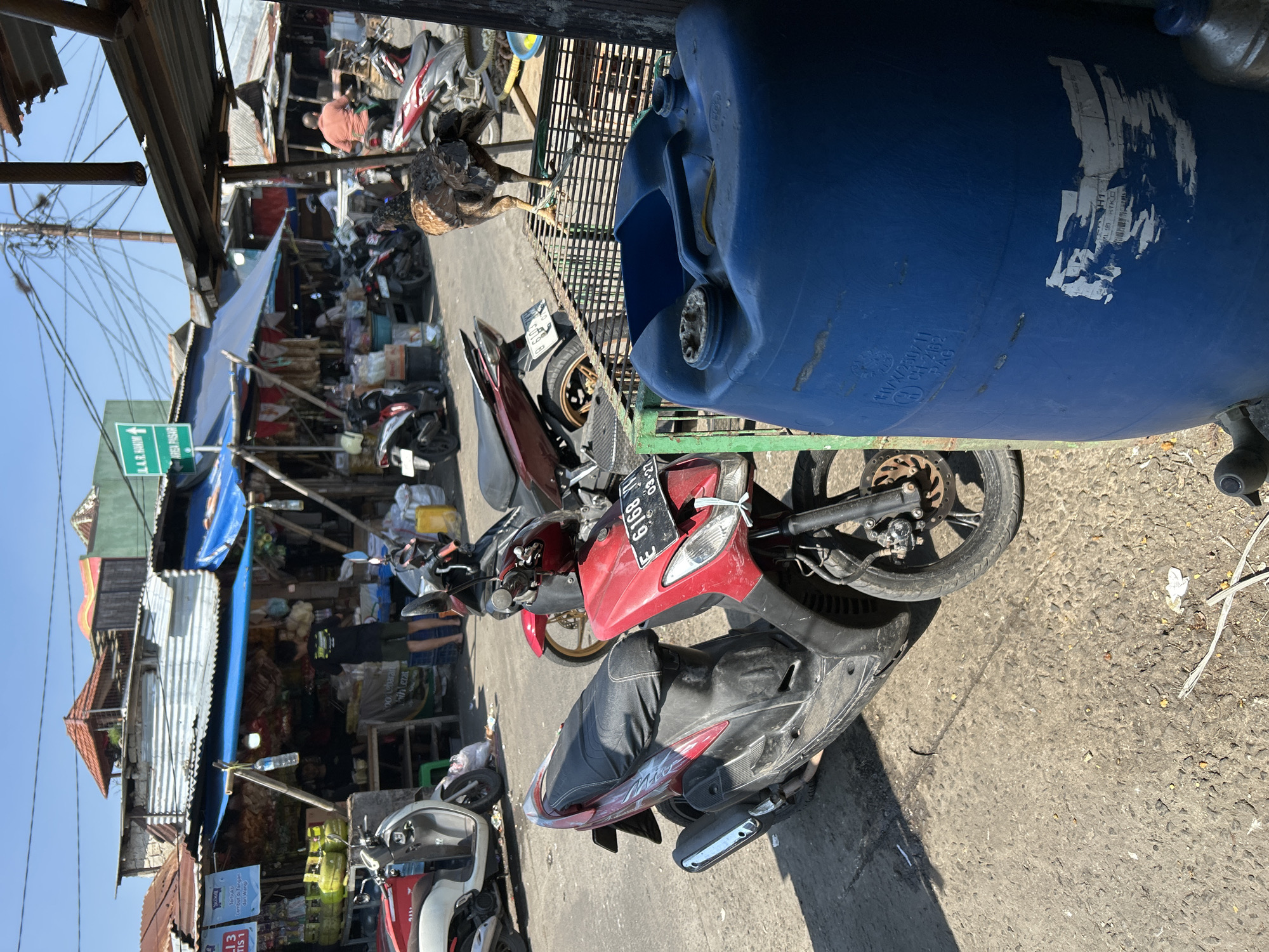 A busy outdoor market scene unfolds on a sunny day in Depok, Indonesia. In the foreground, a red and black scooter is parked on a dusty, asphalt-like surface. To the right, a large, blue plastic barrel stands prominently, partially obscuring the view of the market stalls behind it. A brown and black chicken is perched on a metal grate in front of the barrel.
The market itself is characterized by various stalls with corrugated metal and blue tarp roofs, displaying a variety of goods. Several other motorcycles are visible, parked among the stalls, suggesting active commerce. A few people are present in the market, some appearing to be vendors or shoppers. One person in a black shirt and shorts stands near a stall, while another in an orange shirt is seen walking further into the market. Overhead, a dense network of electrical wires crisscrosses the sky. A green directional sign for "Area Pasar" (Market Area) is visible on a pole in the background. The overall environment appears somewhat unkempt, with debris scattered on the ground.