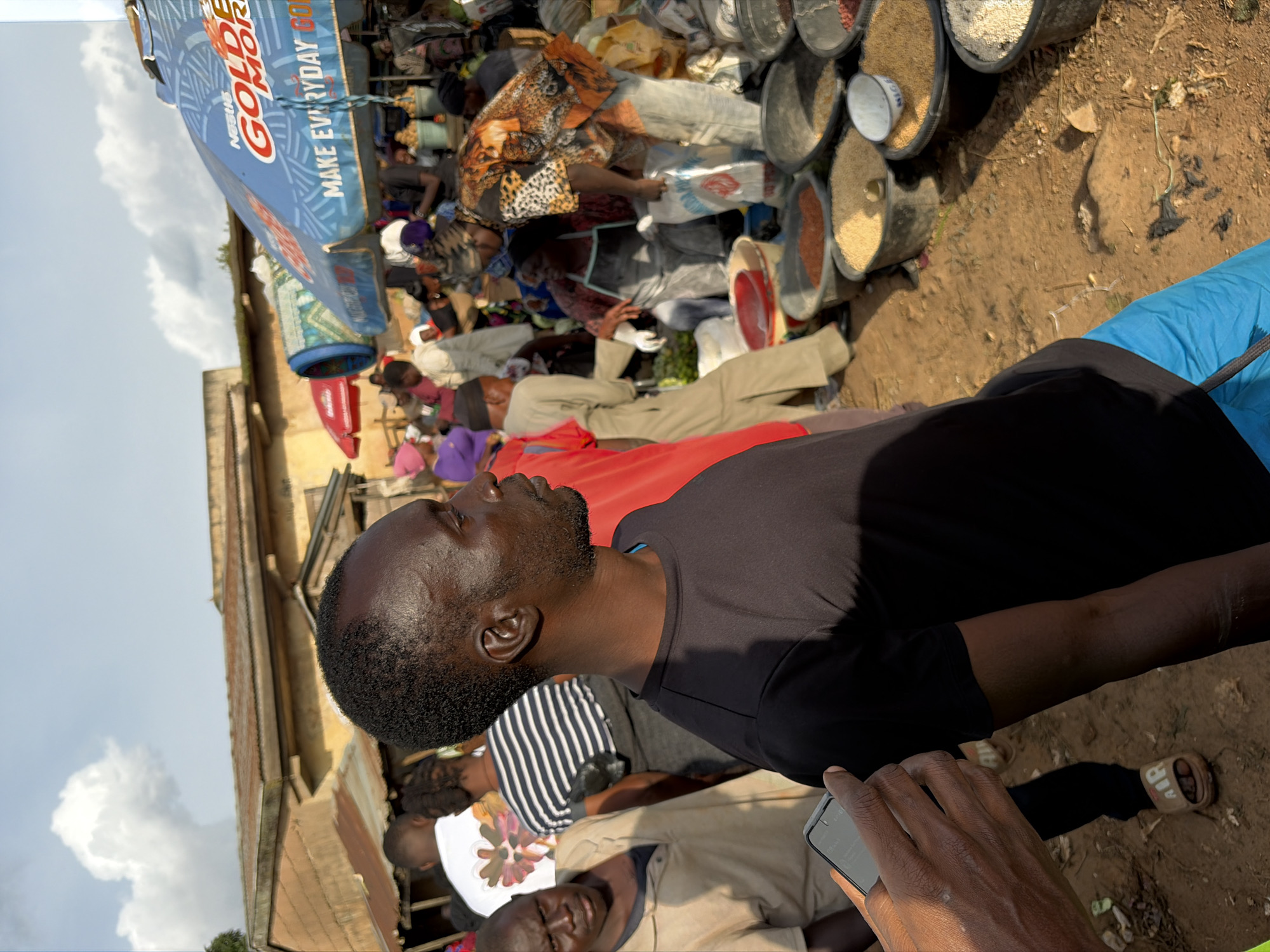 The image depicts a bustling outdoor market scene in Gura Top, Nigeria. A Black man in a black t-shirt and blue shorts is in the foreground, facing right, with his head turned to observe something out of frame. His skin appears dark, and he has short, dark hair. To his left, another Black man in a light-colored polo shirt looks towards the camera with a neutral expression.
Behind them, the market is filled with people engaged in various activities. Stalls are set up with large blue and red umbrellas providing shade. One blue umbrella has the text "NESTLE GOLDEN MORNING" and "MAKE EVERYDAY GOLDEN" printed on it. Various goods are displayed, including large buckets filled with grains and other foodstuffs. Some individuals are seen behind the stalls, possibly vendors, while others are browsing or interacting. The ground is dusty and uneven, characteristic of an outdoor market. The architecture in the background suggests low-rise buildings with corrugated metal roofs. The sky is partly cloudy, indicating daytime. The overall atmosphere is one of lively commercial activity.